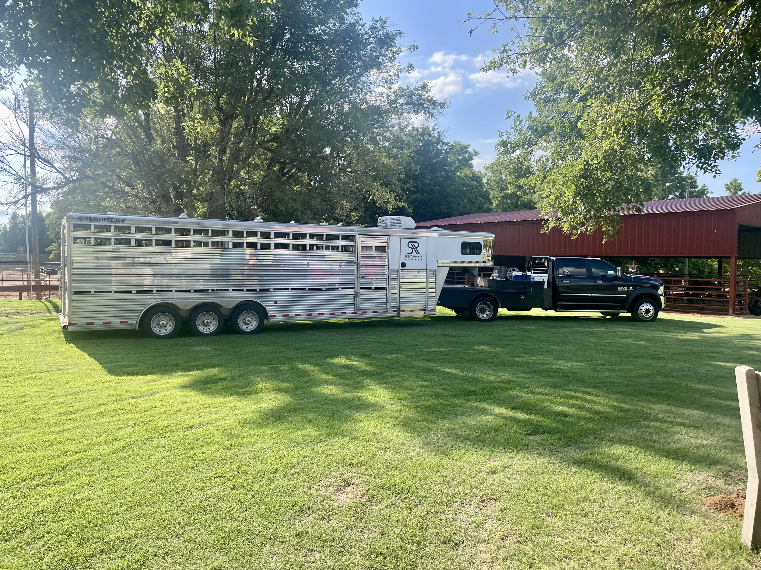 A black pickup truck attached to a large aluminum livestock trailer parked on a grassy area with trees and a red barn in the background. SR Bar Ranch Performance Horses.