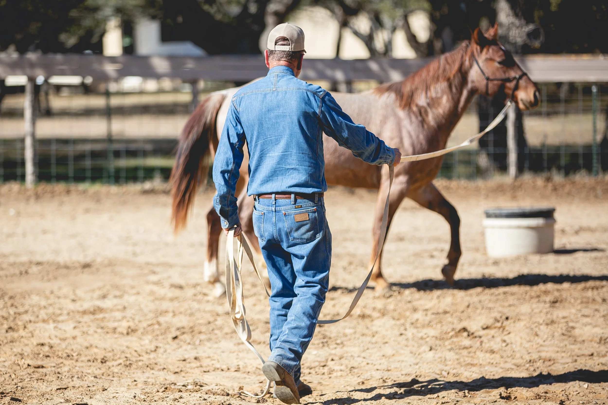 A man wearing a blue denim outfit and a baseball cap leading a brown horse in an outdoor riding area.