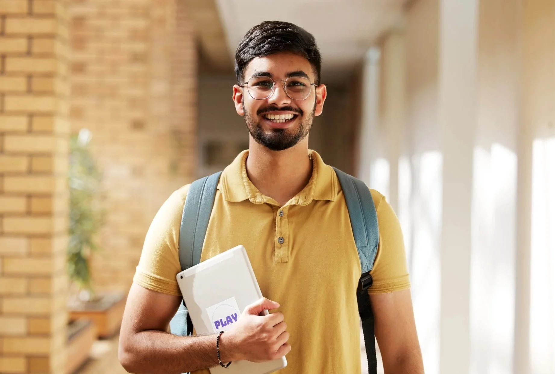 A young man with glasses and a beard smiling, carrying a laptop and wearing a yellow polo shirt and a backpack, standing in a bright hallway with brick walls and large windows.
