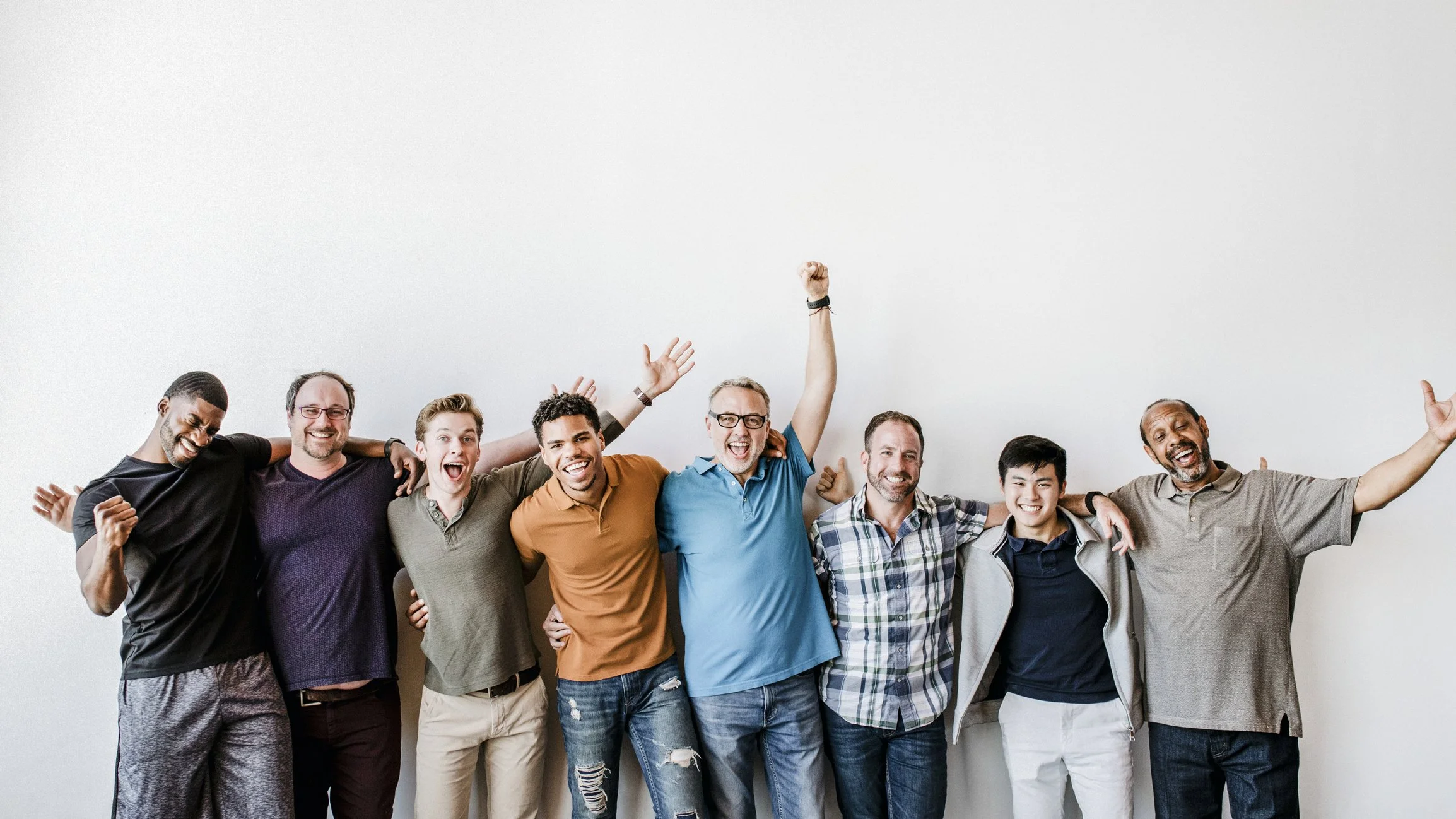A diverse group of nine happy people standing together with their arms around each other, smiling and celebrating against a plain white wall.