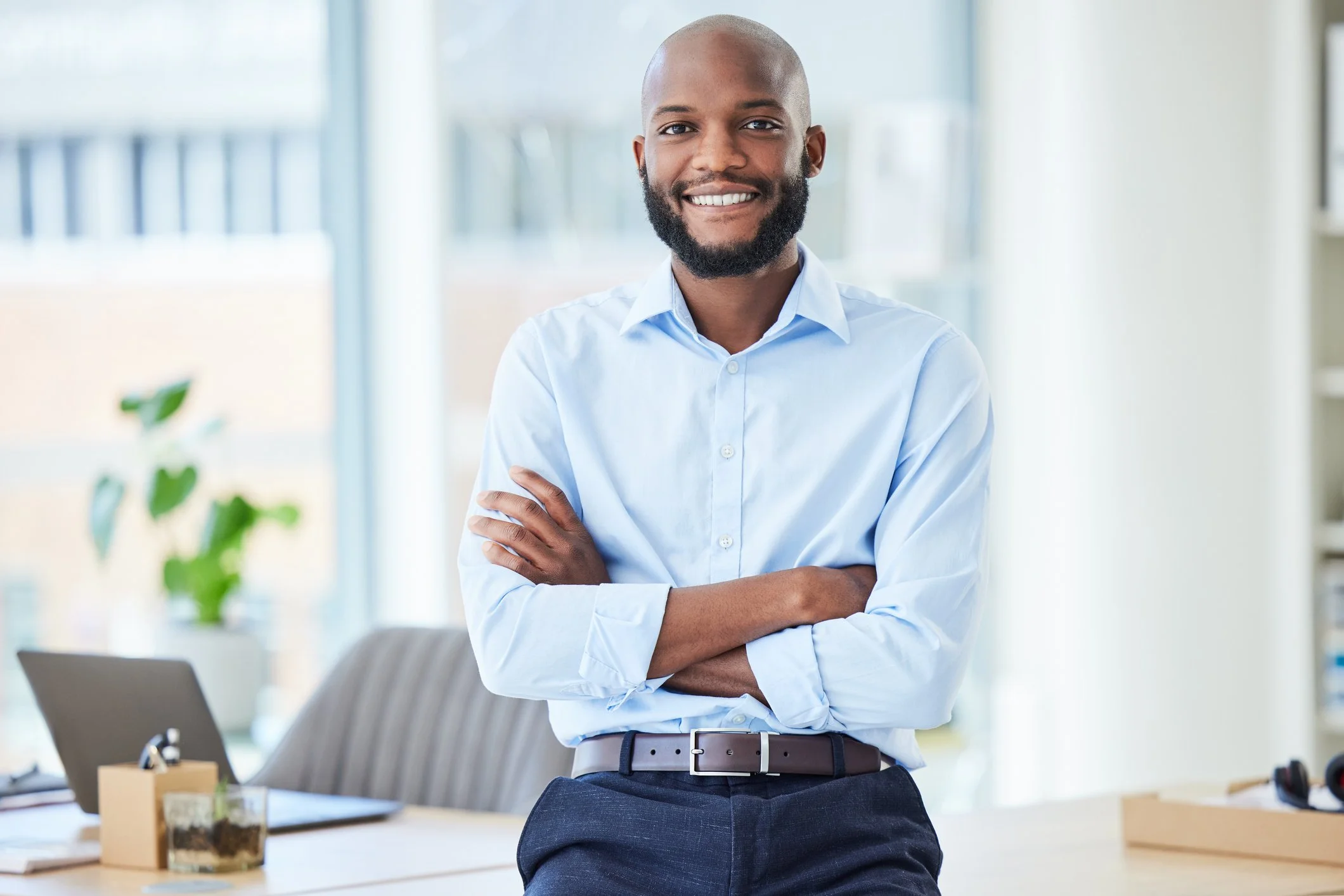 A smiling man with a beard wearing a light blue dress shirt standing with arms crossed in an office space with a desk, plant, and laptop.