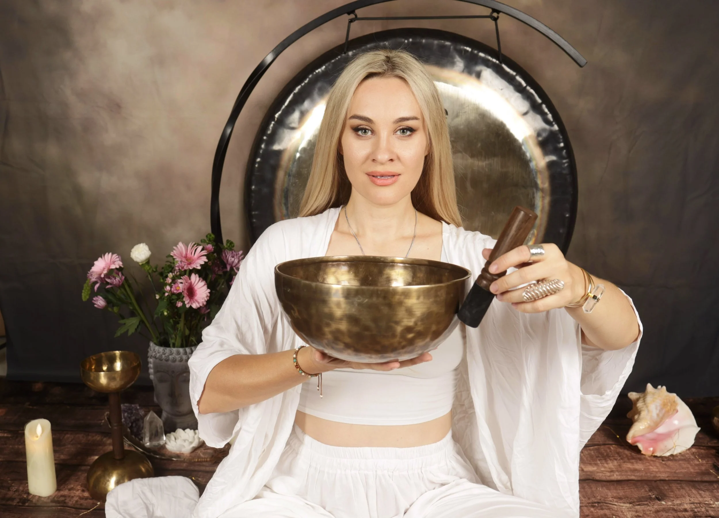 Young woman with long blonde hair holds a large singing bowl, sitting in front of a mirror with candles, flowers, and shells on a wooden table, wearing a white outfit and jewelry.