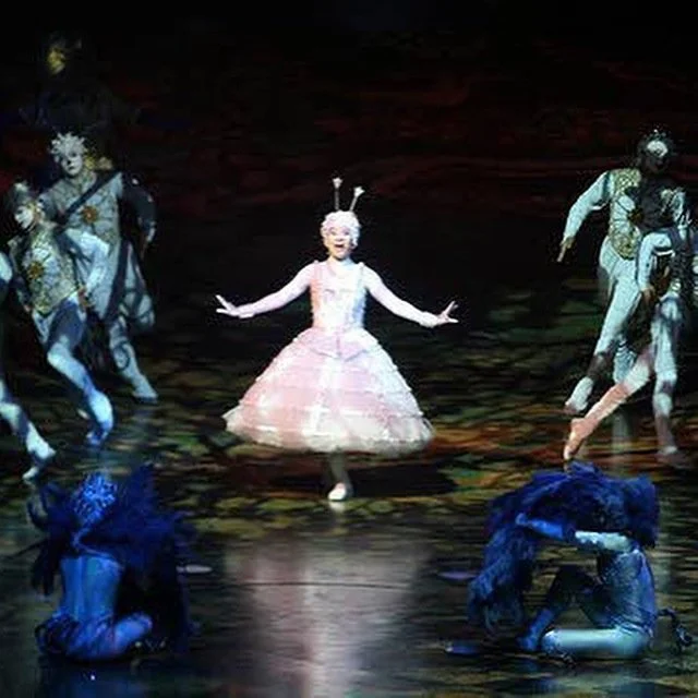 The image shows a stage performance with one performer dressed as a pink insect or ant in the center, surrounded by other performers dressed in elaborate costumes, some in blue and others in black and white. The scene is colorful with dim stage lighting.