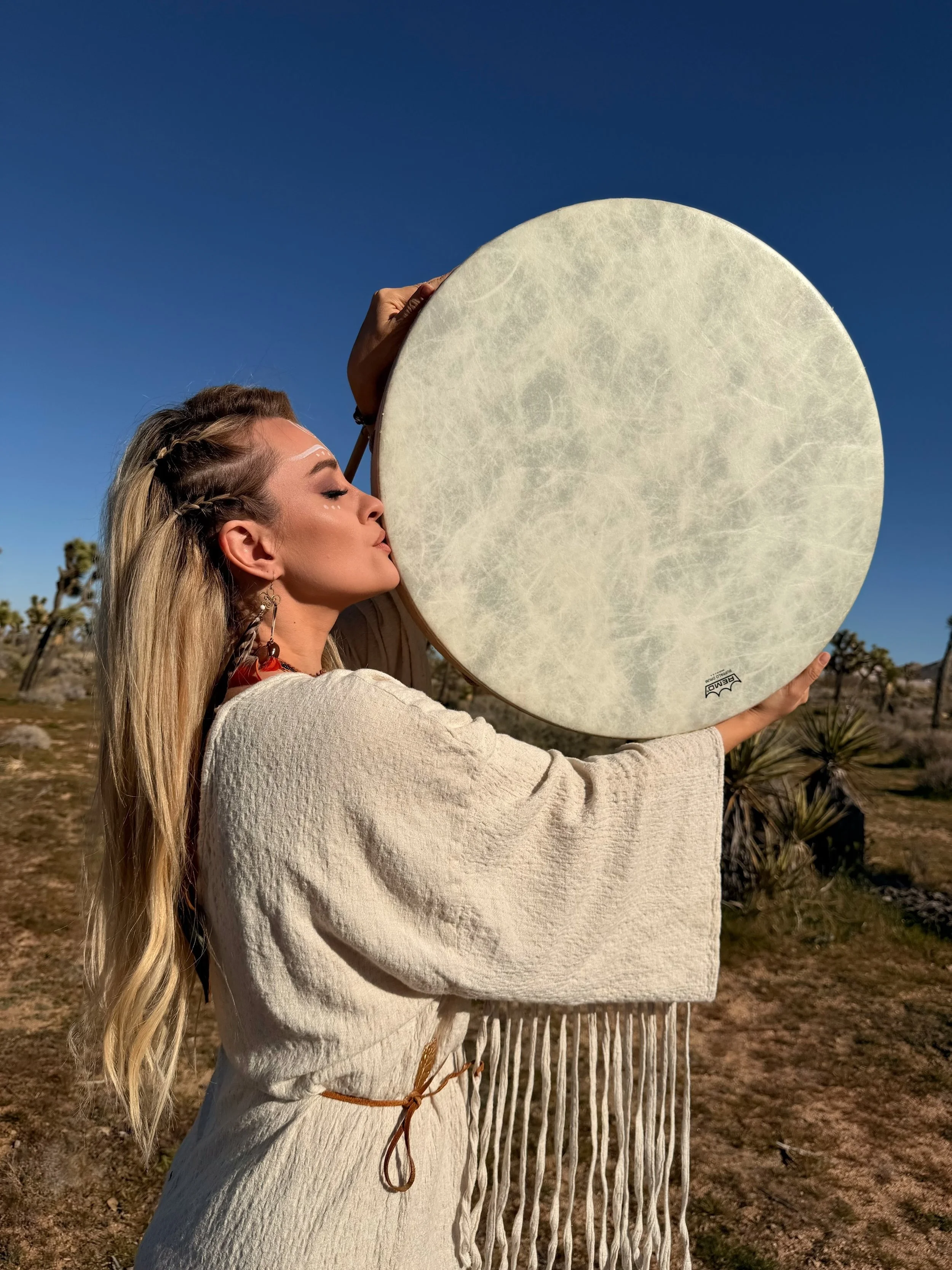 A woman in a cream-colored dress with fringe details holds a large round drum close to her face in a desert landscape with Joshua trees under a clear blue sky.