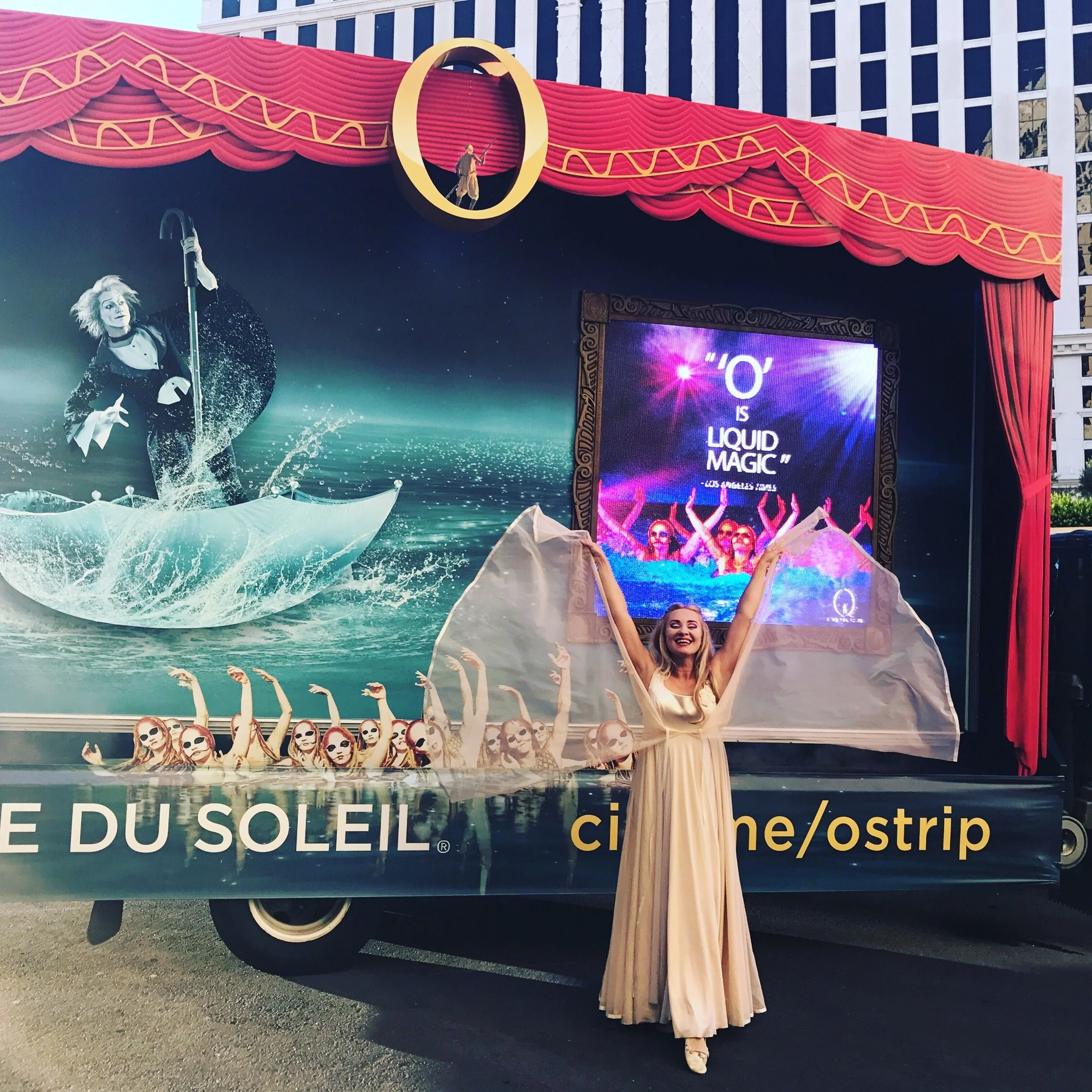 A woman in a long beige dress with sheer fabric on her arms, standing in front of a movie-themed float with circus/film festival theme, smiling with arms raised. The float features a large screen with a quote about liquid magic, a stylized woman in a black dress and white hair, and multiple women in sunglasses in a water scene, with a backdrop of a starry sky and ocean.