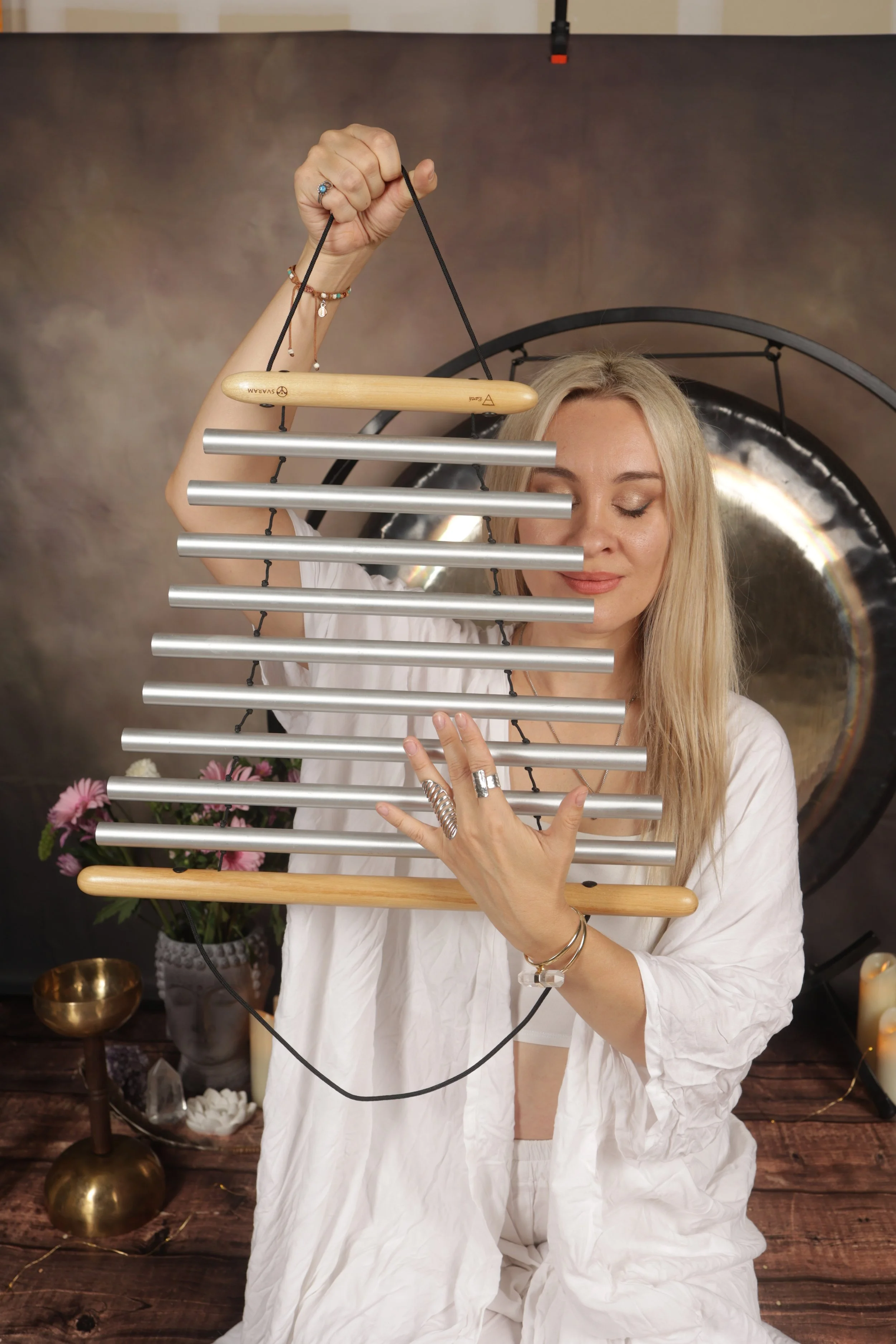 A woman with long blonde hair and closed eyes playing a metal and wooden musical instrument, possibly a wind chime or bell set, in a room with a gong, candles, and flowers in the background.