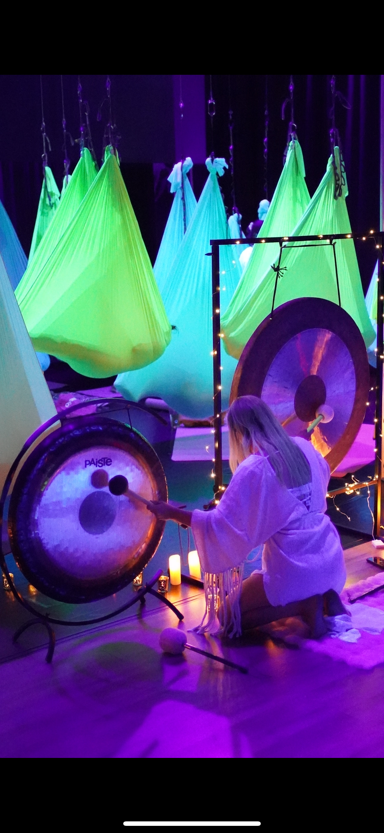Person kneeling and playing a gong in a dimly lit room with hanging fabric structures illuminated in neon green, purple, and blue colors, decorated with fairy lights and candles.