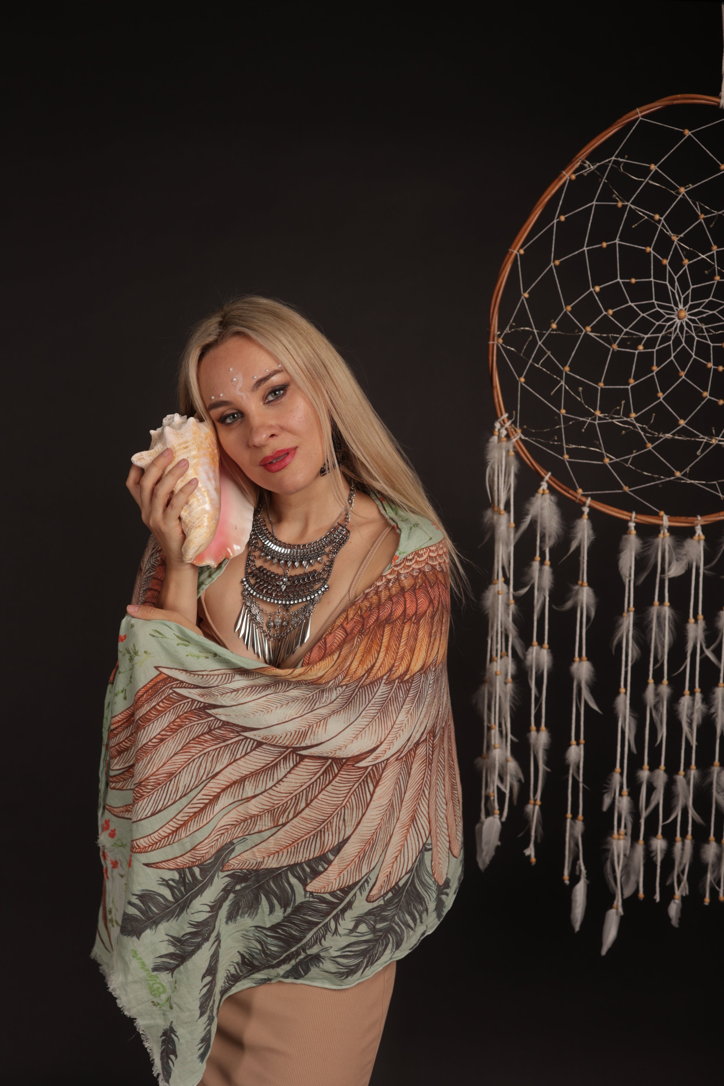 A woman with blonde hair holding a large seashell to her face, wearing layered necklaces and a feather-patterned shawl, standing next to a large dreamcatcher against a dark background.