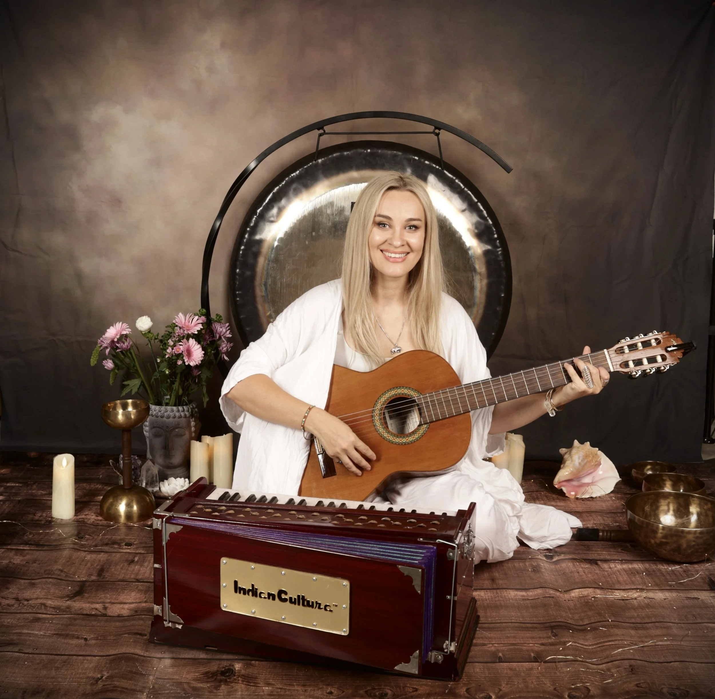 A woman with long blonde hair wearing a white dress playing an acoustic guitar in a decorated setting with a keyboard, candles, flowers, and a gong behind her.