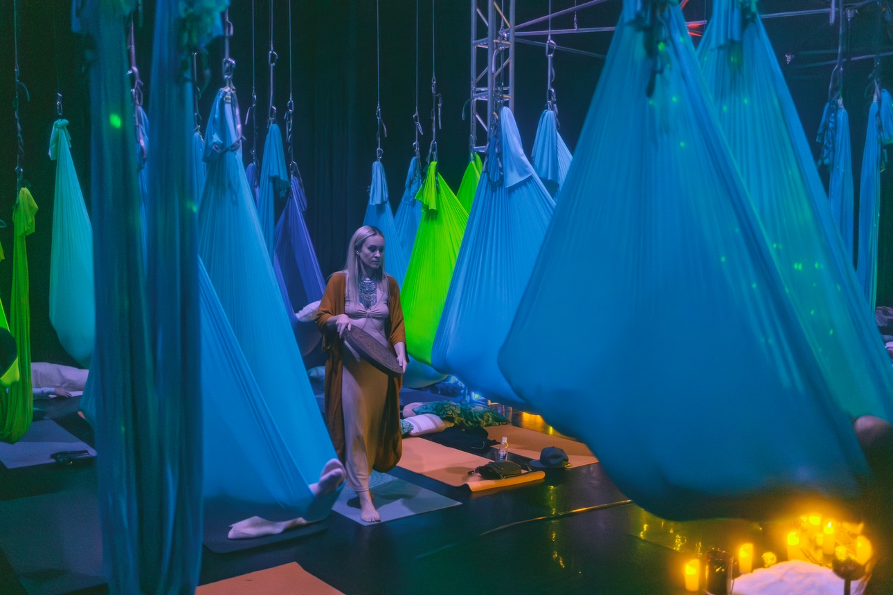 A woman in a long dress and cardigan standing among suspended blue and green fabric hammocks in a dark room illuminated by yellow candles, possibly a yoga or relaxation studio.