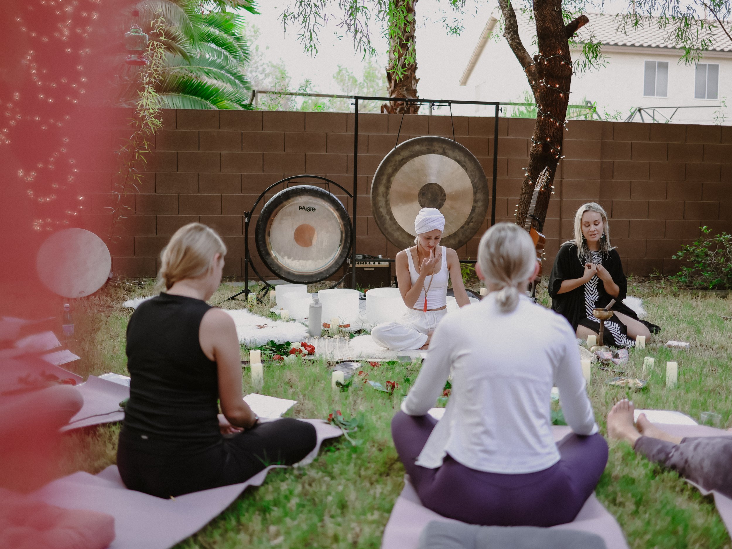 Group of women participating in a meditation or sound healing session outdoors in a backyard, with gongs, singing bowls, candles, and feng shui elements around them.