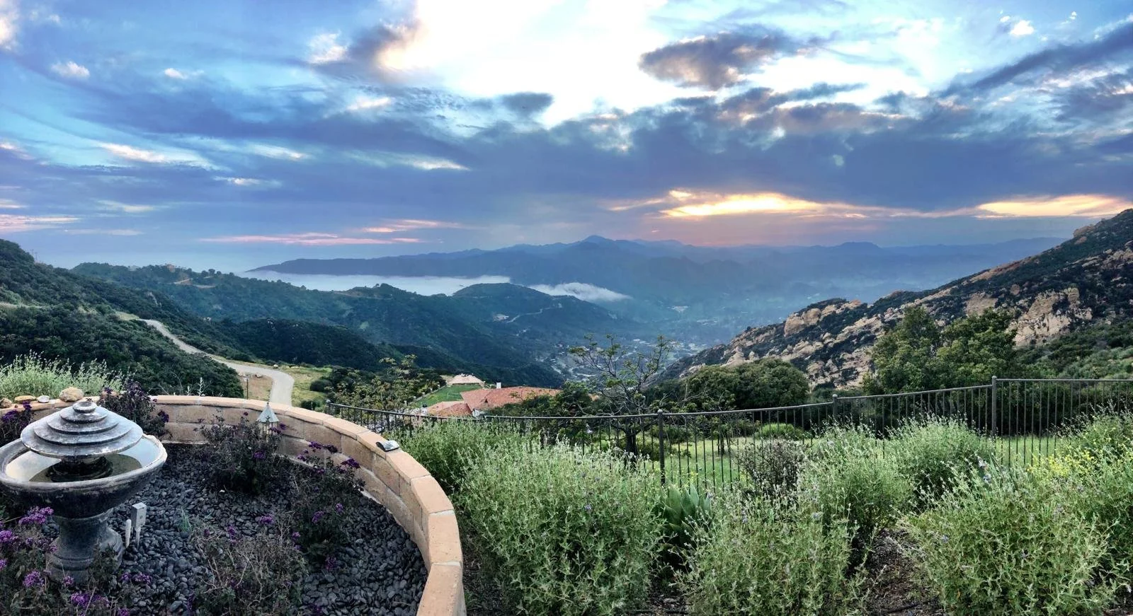 Scenic view of a lush green valley with mountains in the distance during sunset, hills covered with trees, and a garden area with plants and a decorative fountain in the foreground.