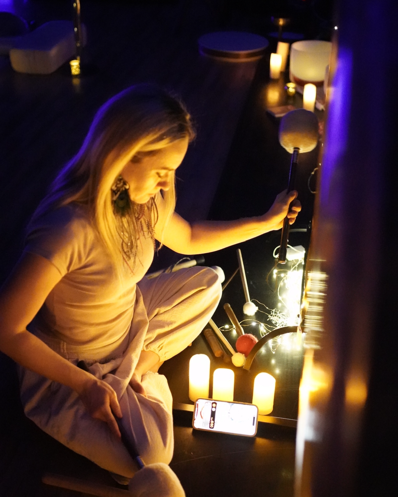 A woman sitting cross-legged on the floor in a dimly lit room, surrounded by candles, holding a mallet, with a smartphone placed in front of her taking a mirror selfie.