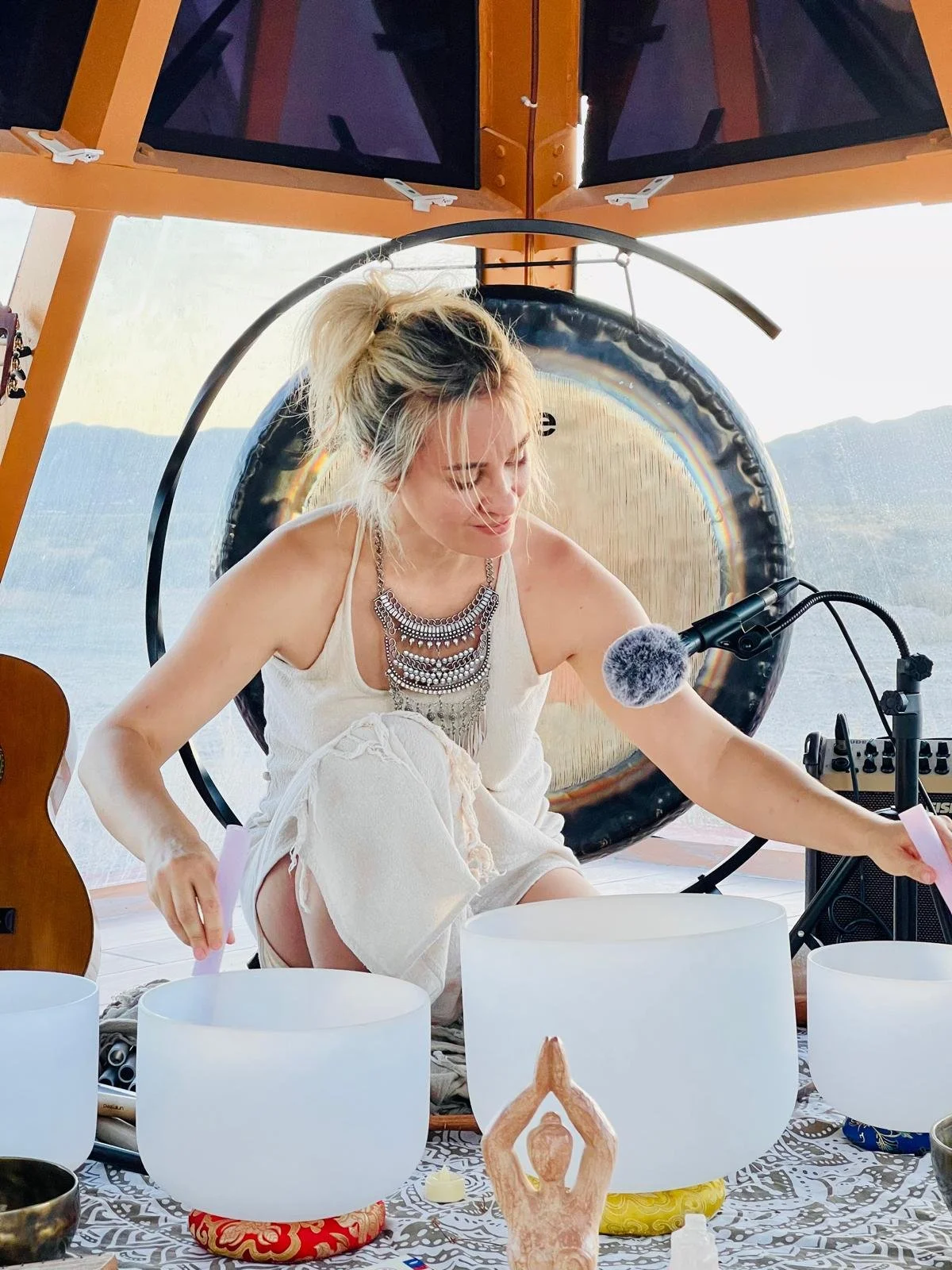 A woman with blonde hair in a messy bun, wearing a sleeveless white top and patterned jewelry, is sitting on the floor and playing crystal singing bowls in a room with a mountain view through large windows. She has a microphone positioned near her mouth and appears focused on her activity.