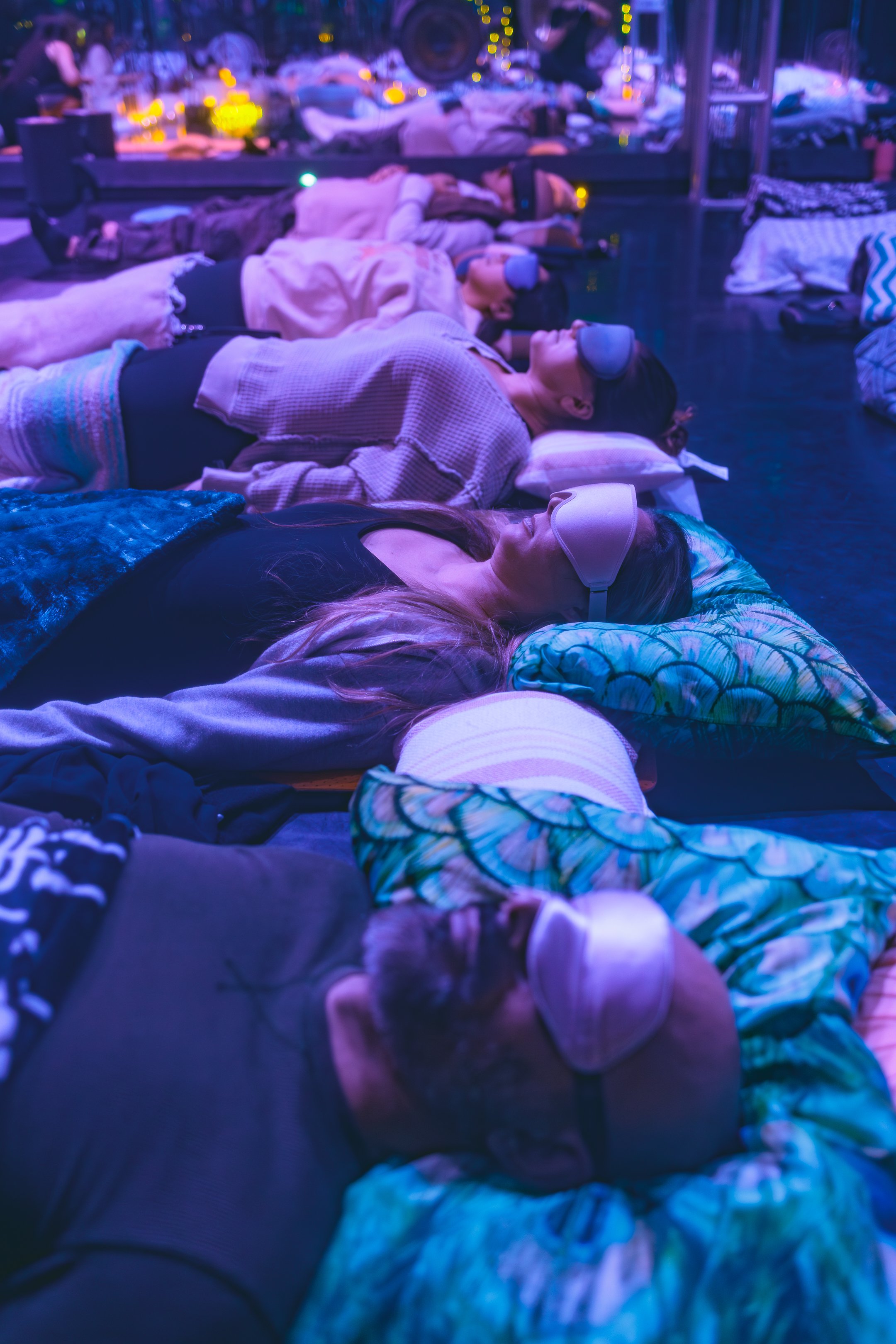 People lying on mattresses with pillows in a dark room, wearing sleep masks, engaging in a relaxation or sleep activity.