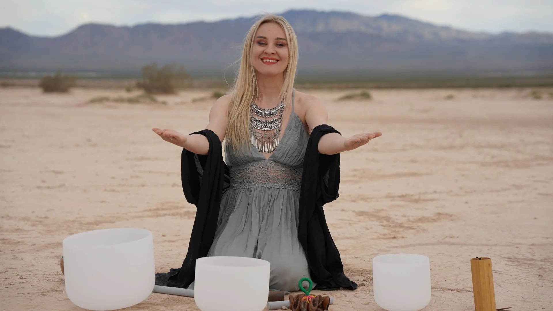 A woman with long blonde hair, wearing a grey dress and layered silver jewelry, sits on the desert ground with her arms outstretched, surrounded by musical instruments including crystal singing bowls and a wooden percussion instrument, in a desert with mountains in the background.