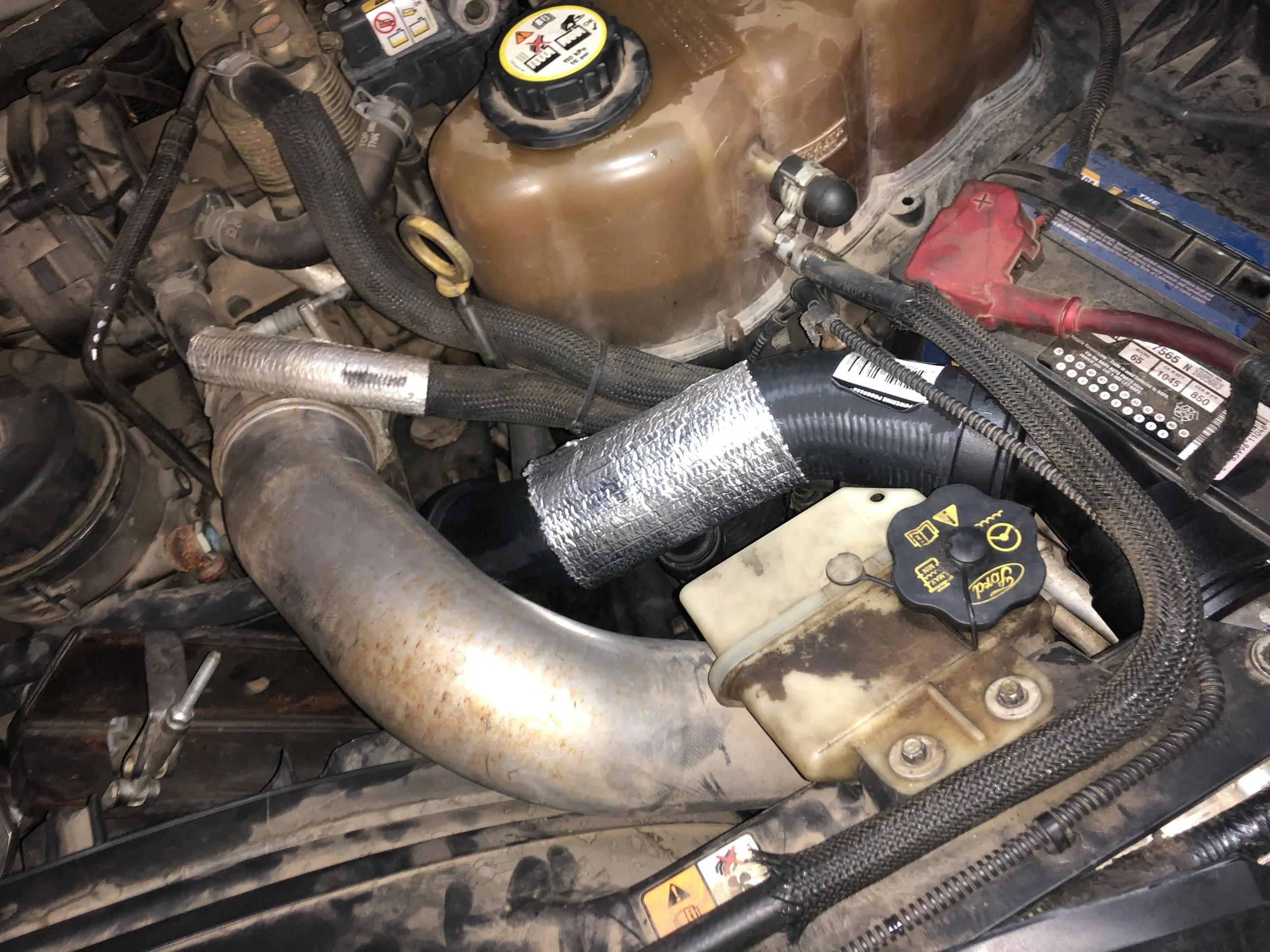 Close-up of a car engine compartment showing hoses, a radiator cap, a battery, and various engine components.