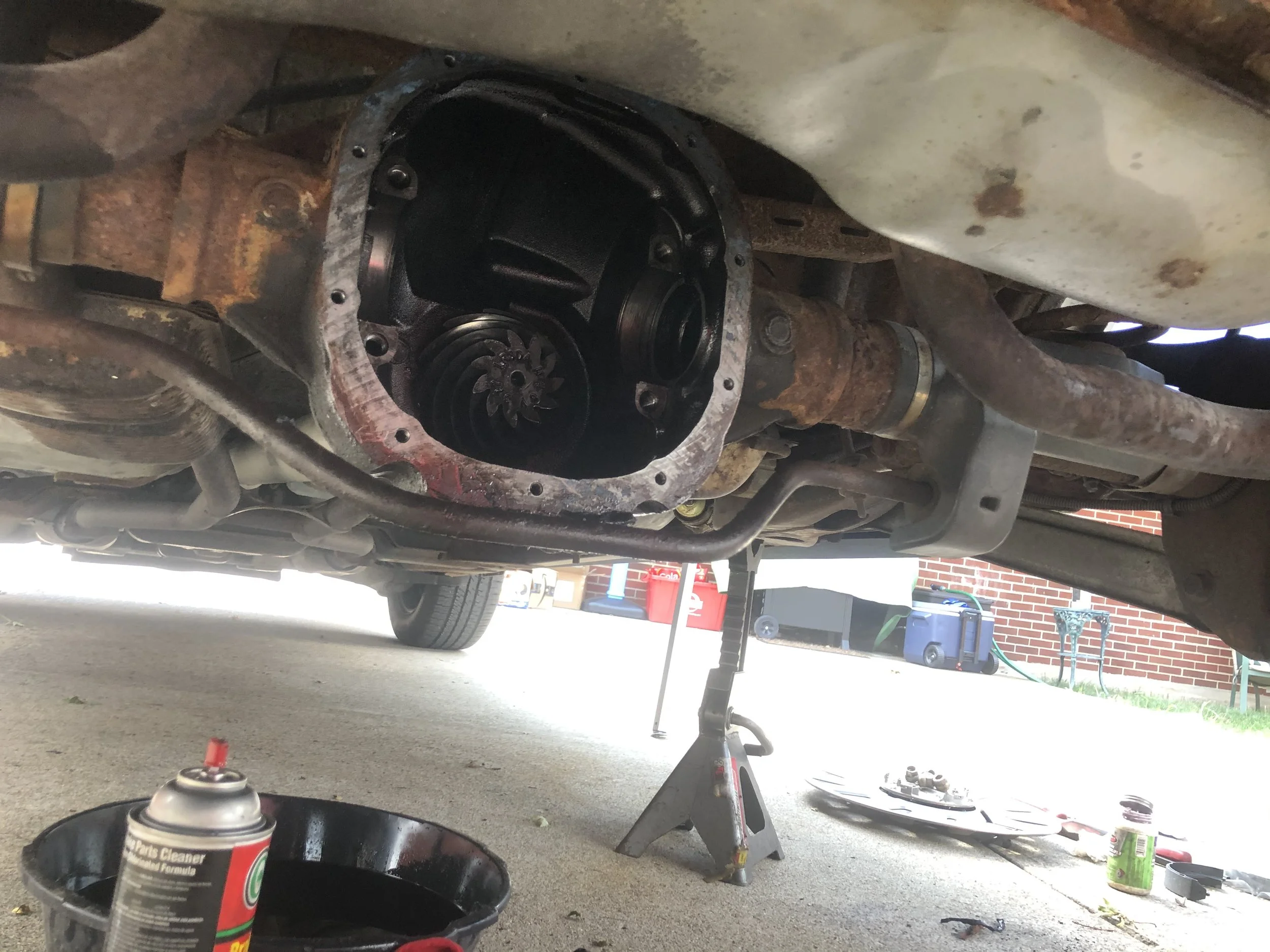 Underneath a vehicle with the oil pan removed, exposing the internal parts. The vehicle is supported by a jack stand, and various tools and parts are on the ground nearby.
