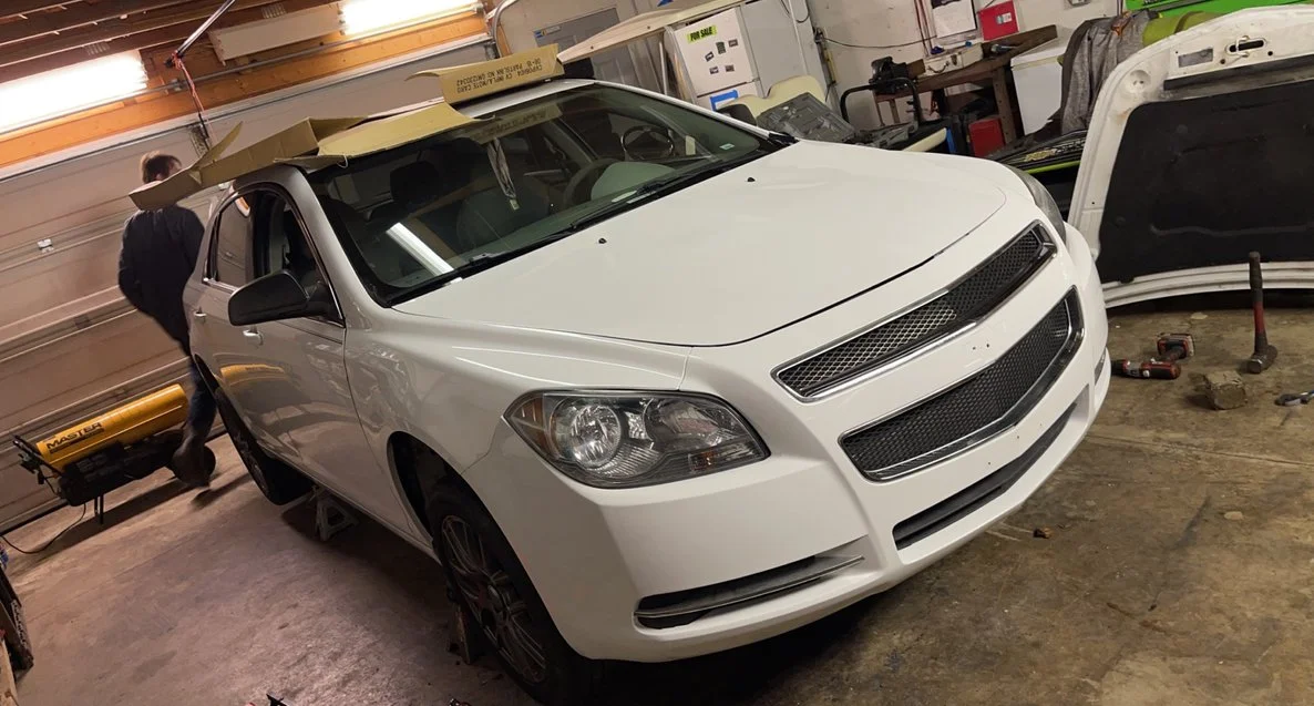 A white sedan inside a garage with a person standing next to it. The garage has tools and car parts, including a car hood and a portable heater.