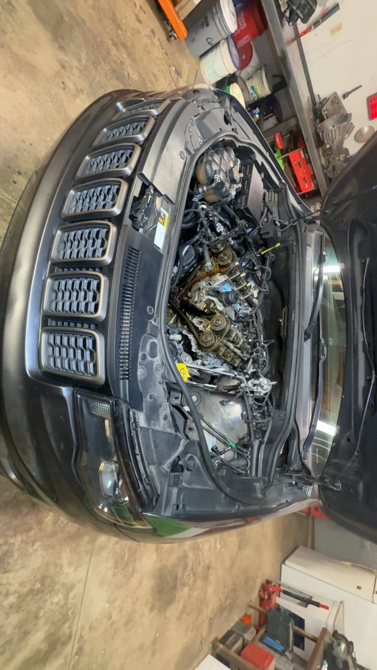 Front of a car with the hood open showing the engine, in a garage with shelves and paint buckets in the background.
