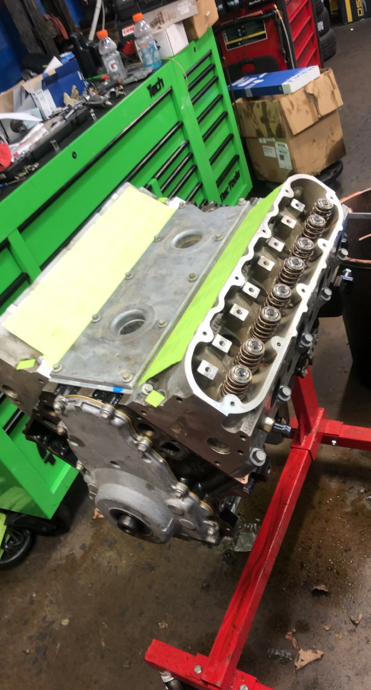 An opened engine head on a red engine stand in a workshop, with a green tool chest, cardboard boxes, and workshop supplies in the background.