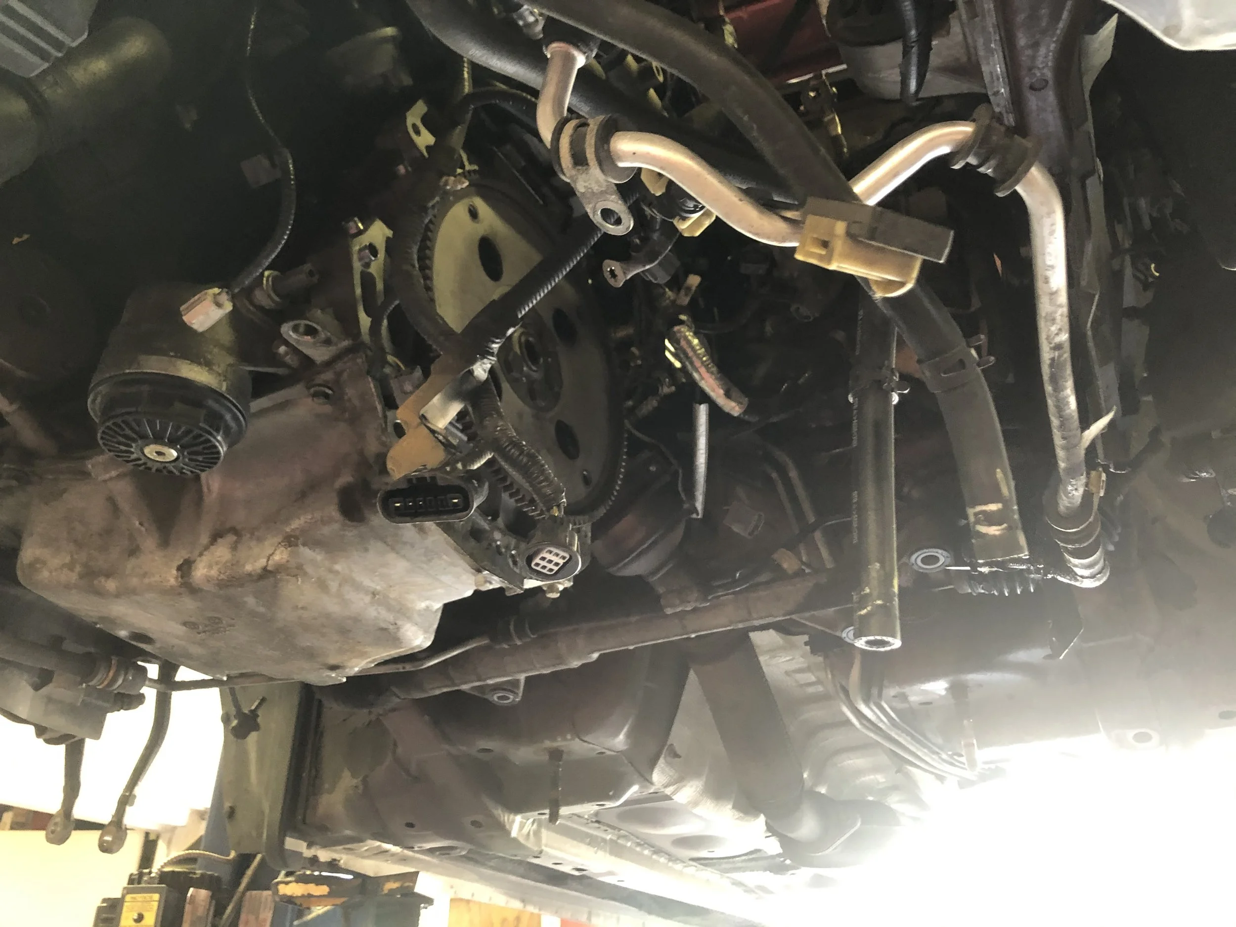 Underneath view of a vehicle engine with visible belts, hoses, and connectors.