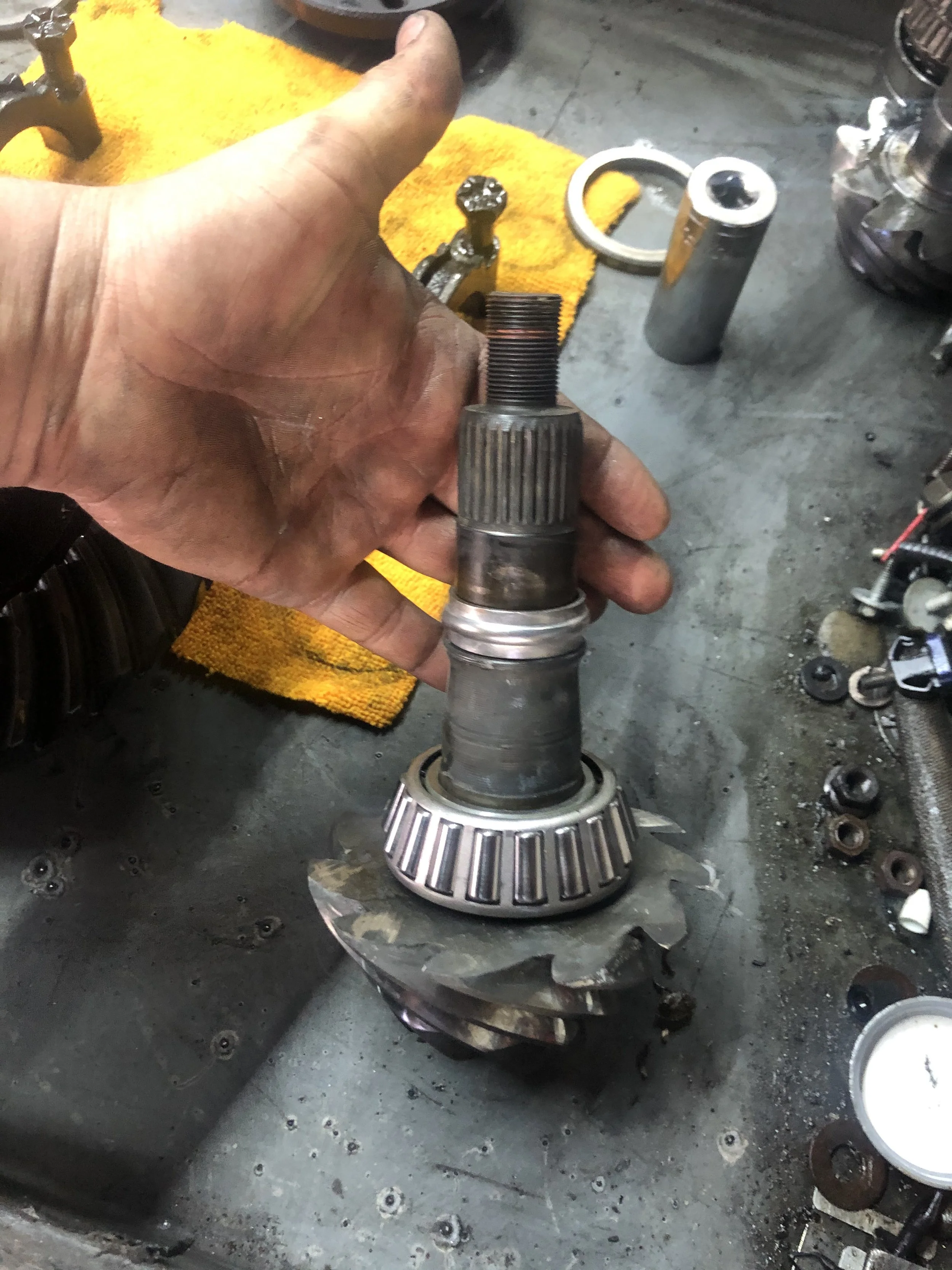 Close-up of a hand holding a transmission gear shaft inside a car repair shop with various tools and parts on the workbench.