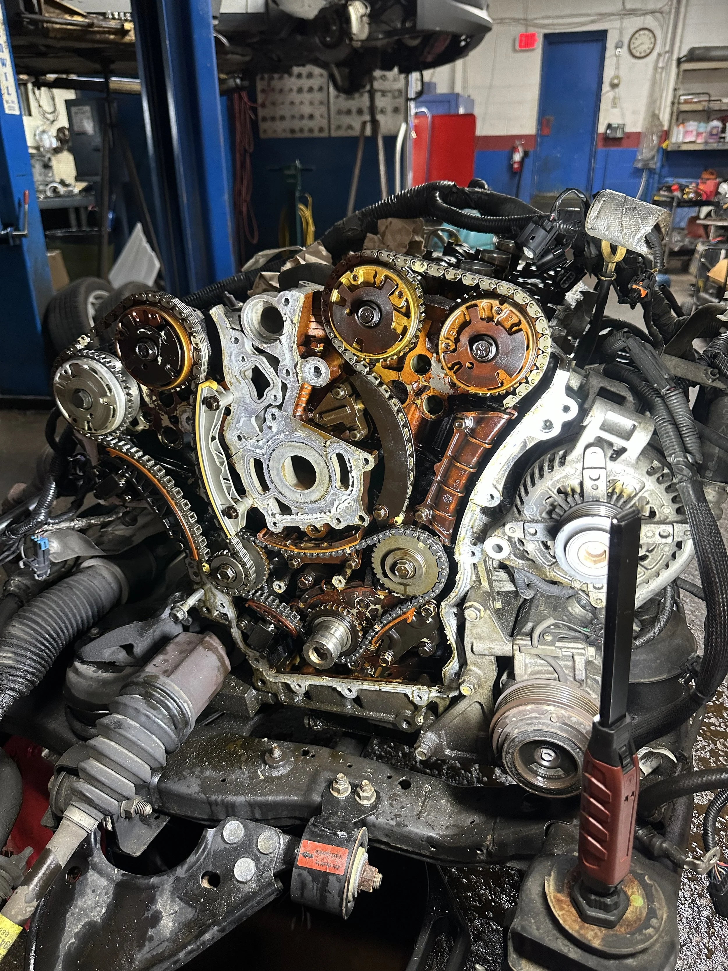 Close-up of a car engine with timing chain and gears exposed, inside an automotive repair shop.