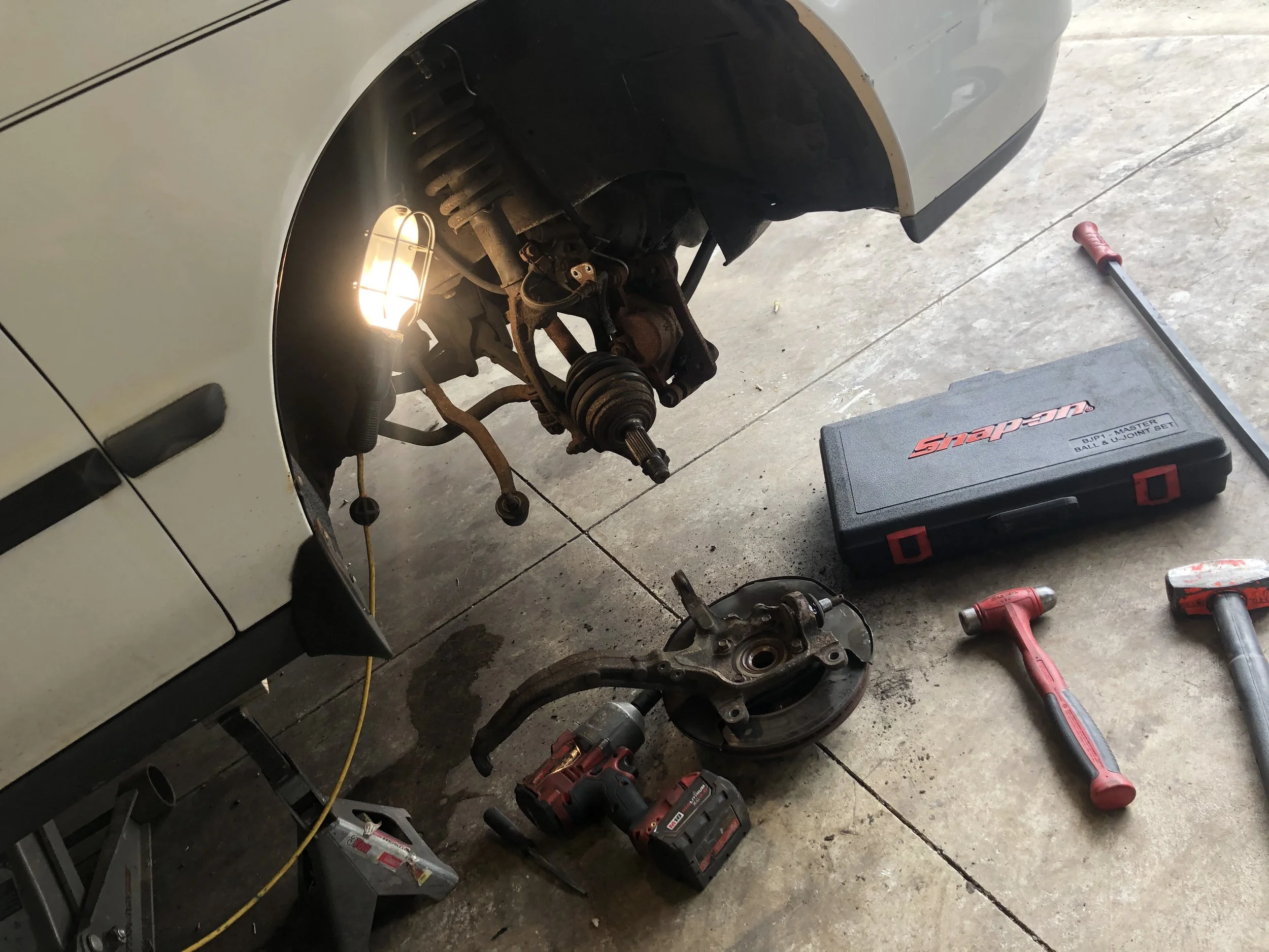 A car with front wheel removed, exposing suspension and brake components, set on a garage floor. Tools including a cordless drill, a rubber mallet, a hammer, and a tool case are arranged nearby.