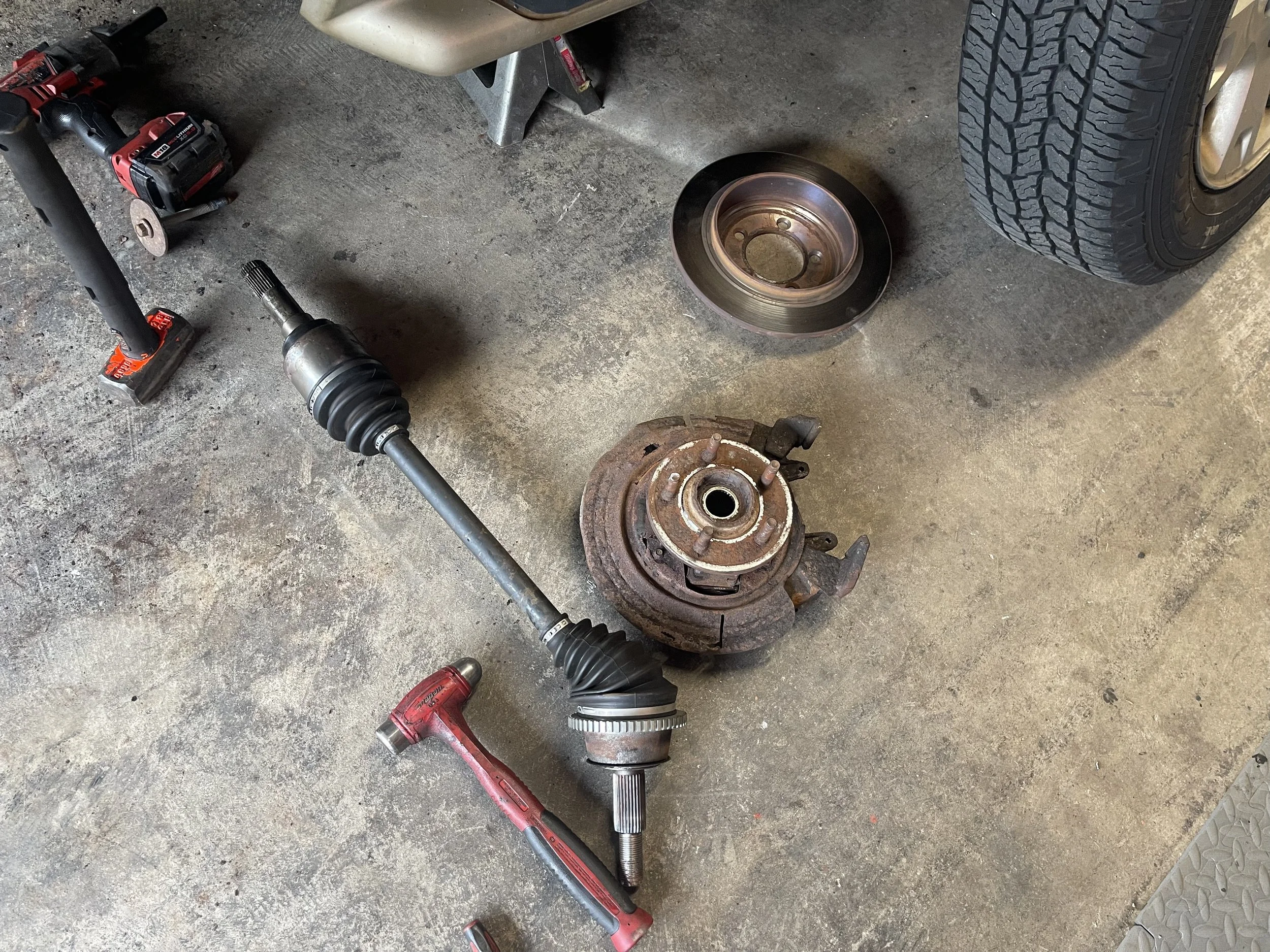 Disassembled car wheel hub, rotor, and CV axle on garage floor, with tools and spare tire nearby.