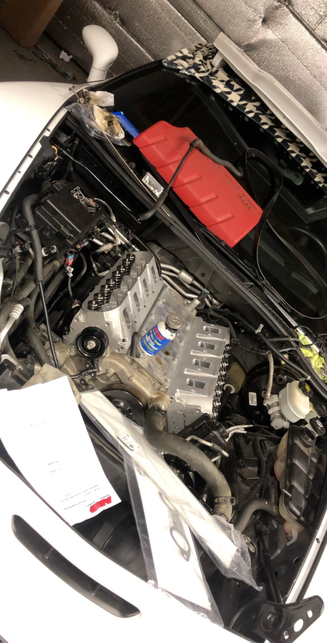 Car engine with hood open, showing engine components and tools, including a red fuel container in the background.