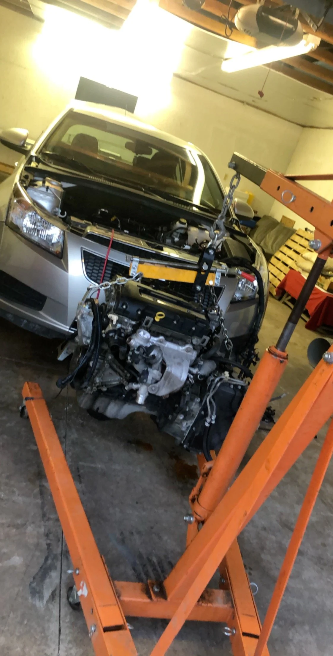 A car with its engine removed is in a garage, lifted by an orange engine hoist.