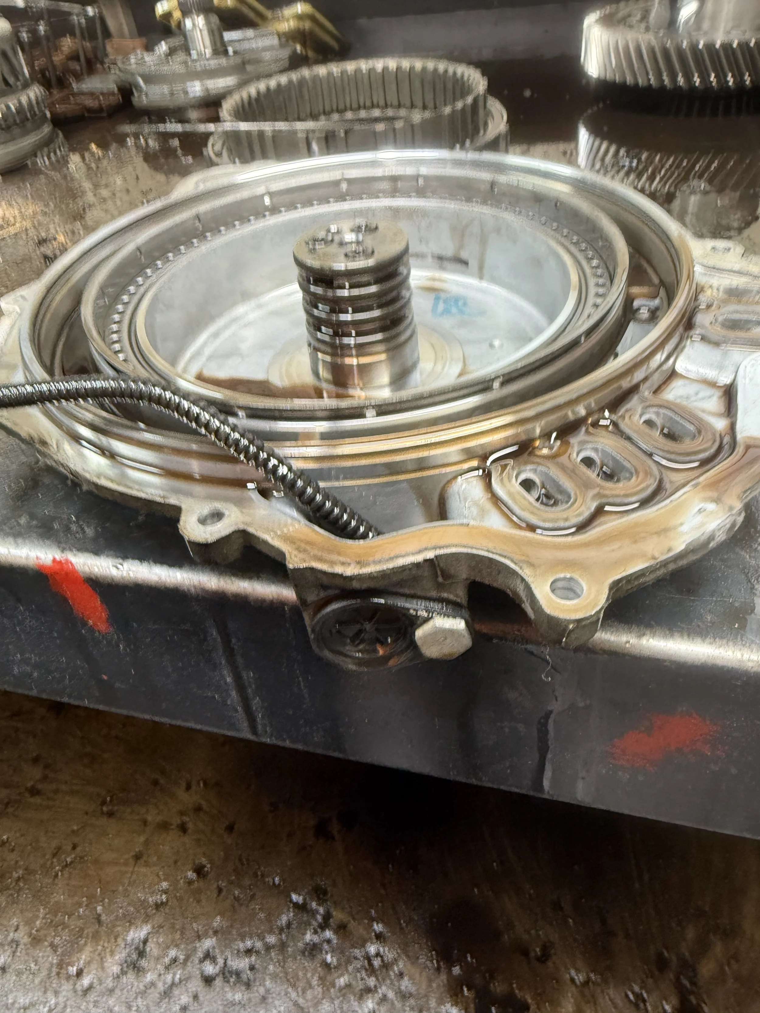 Close-up of a disassembled engine component, showing a large circular metal part with a central shaft and several gaskets, placed on a workbench with visible rust and dirt.