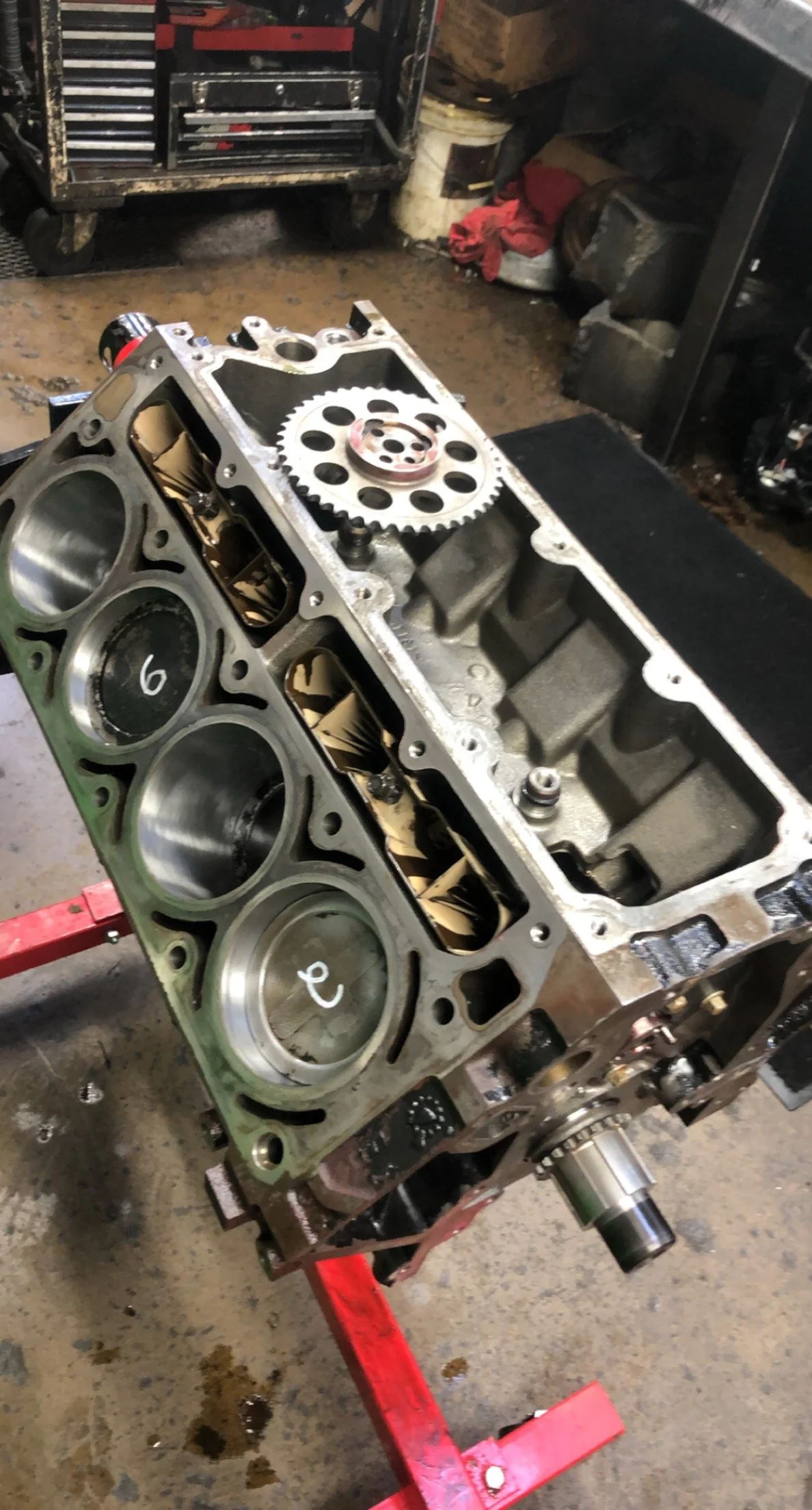 A partially assembled engine block on a red engine stand with pistons, valves, and gears inside a workshop.