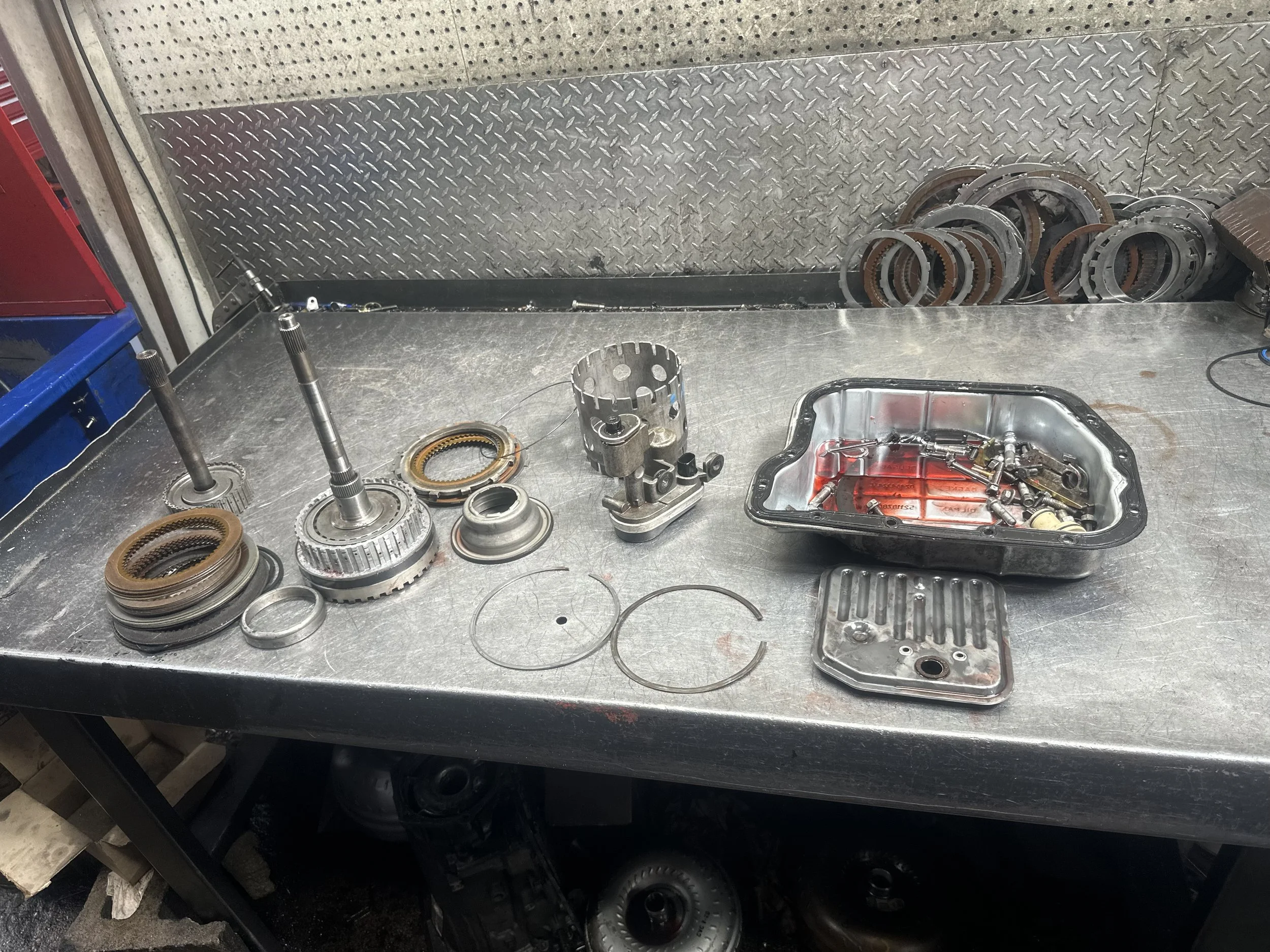 Disassembled automotive transmission components on a metal workbench, including gears, bearings, a valve body, and a transmission pan with red fluid.