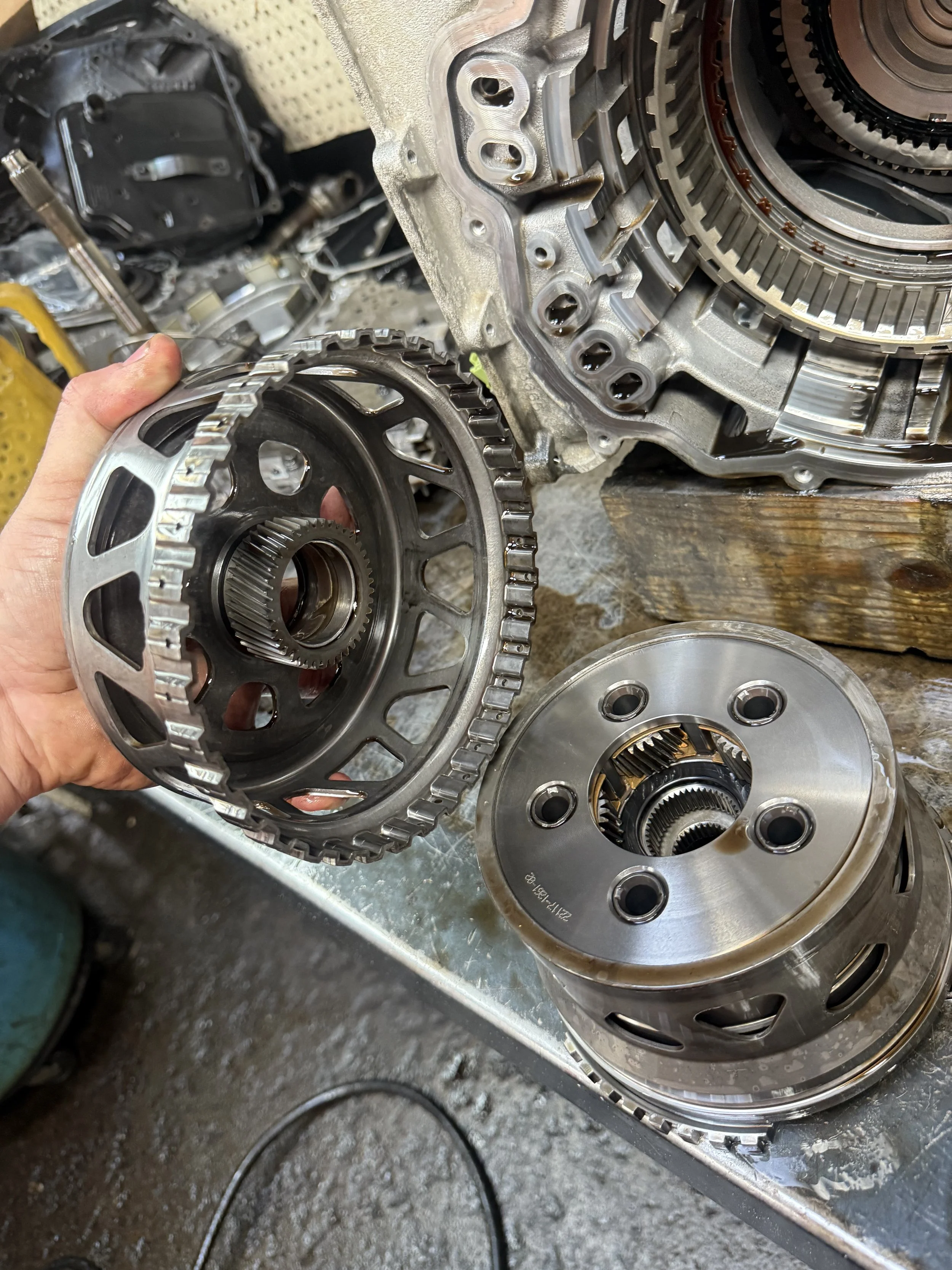 Close-up of a person's hand holding a gear assembly next to another gear assembly, with an engine or transmission housing in the background on a workbench.
