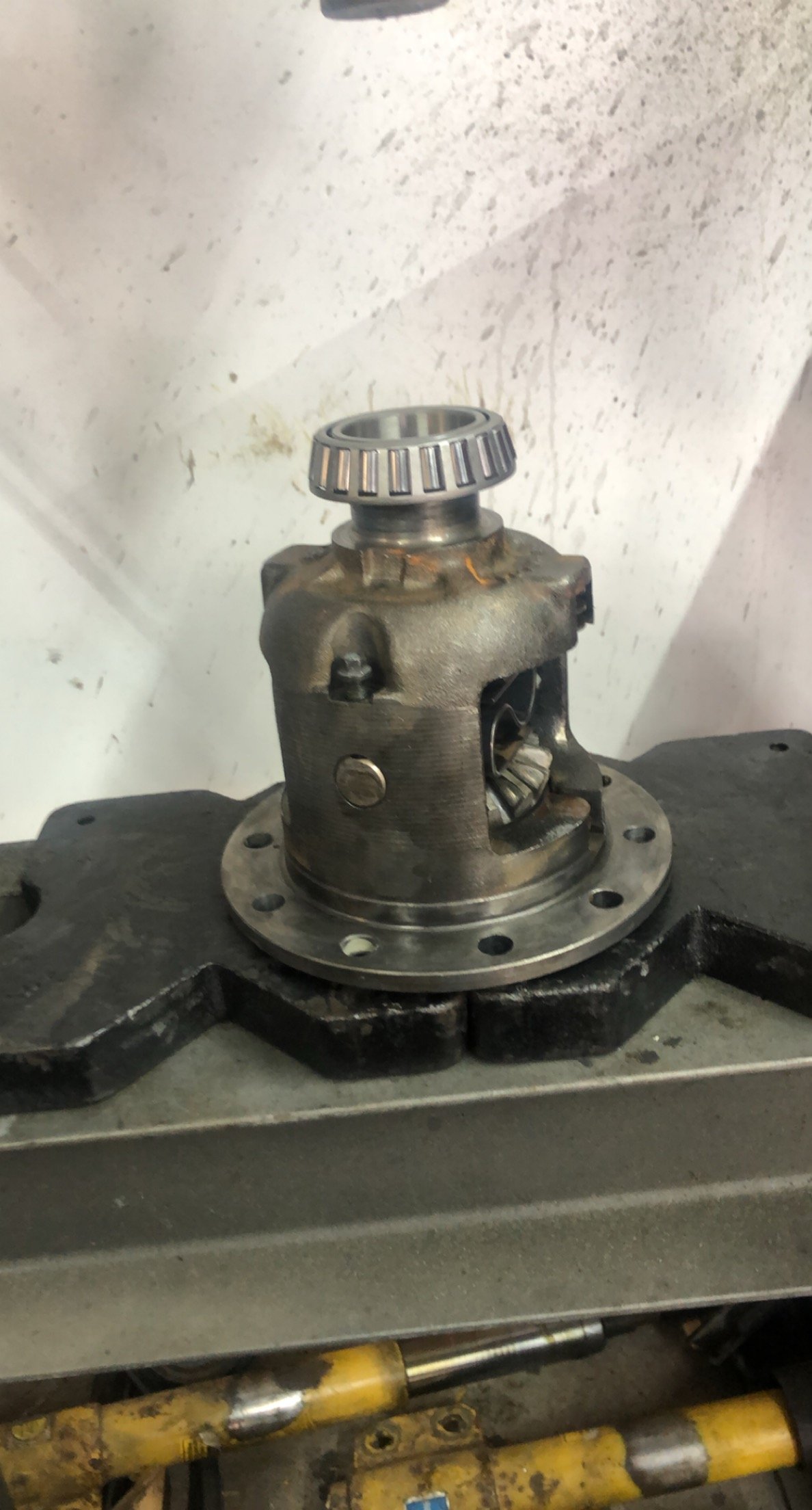 A mechanical component, possibly part of a machine or vehicle, placed on a work surface. It is metallic with a flange base and a cylindrical body, featuring an opening and gear-like structures at the top.