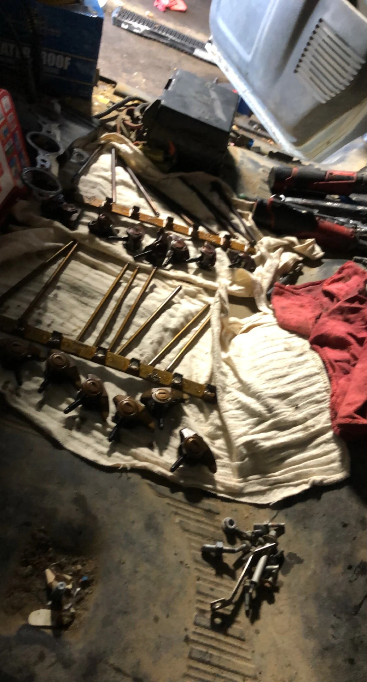 Various automotive parts and tools laid out on a cloth, including shock absorbers, control arms, and sockets, with a workbench and other workshop items in the background.