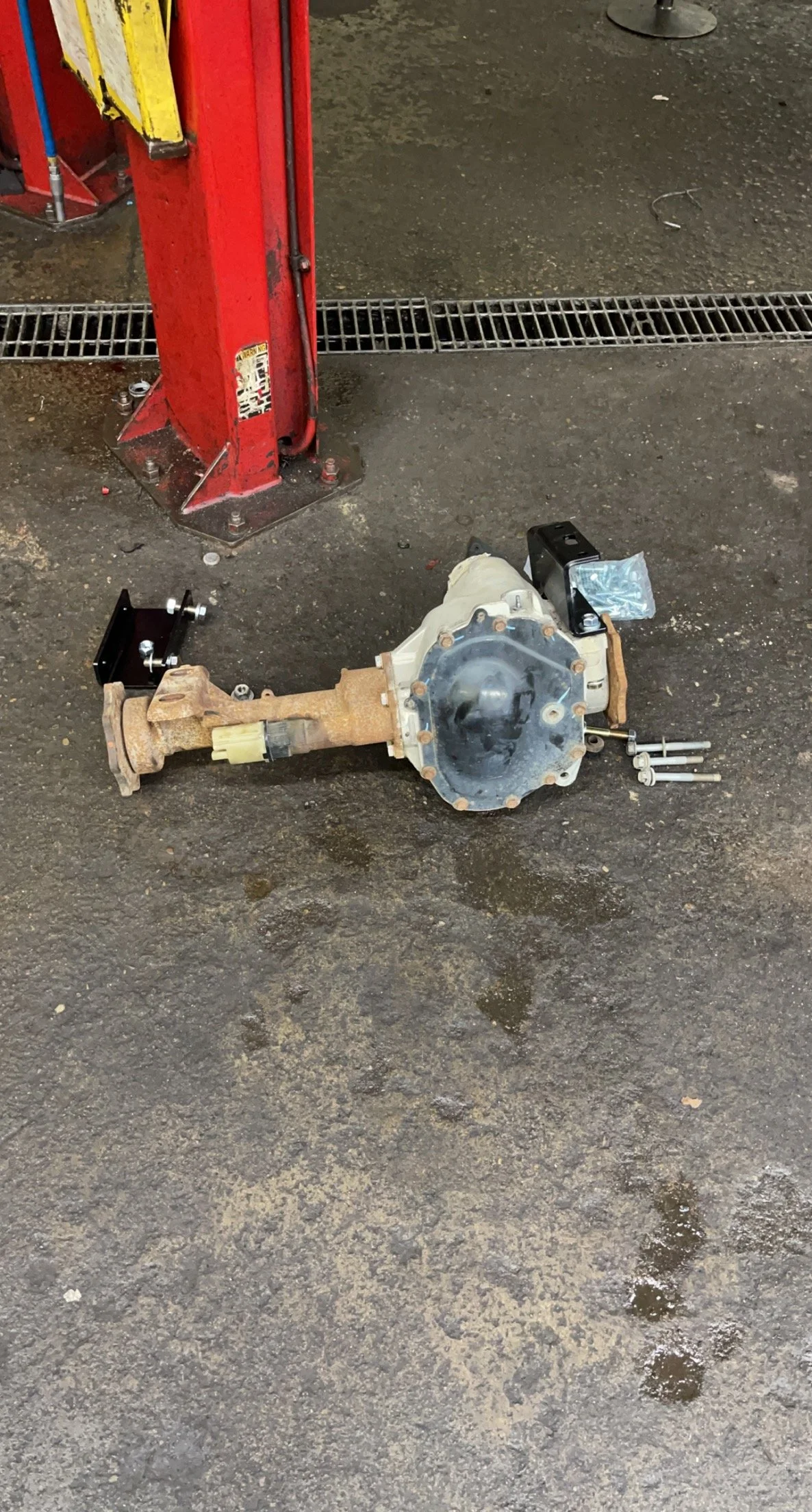 Automobile differential axle assembly placed on a concrete workshop floor with a red hydraulic lift and a drain grate nearby.