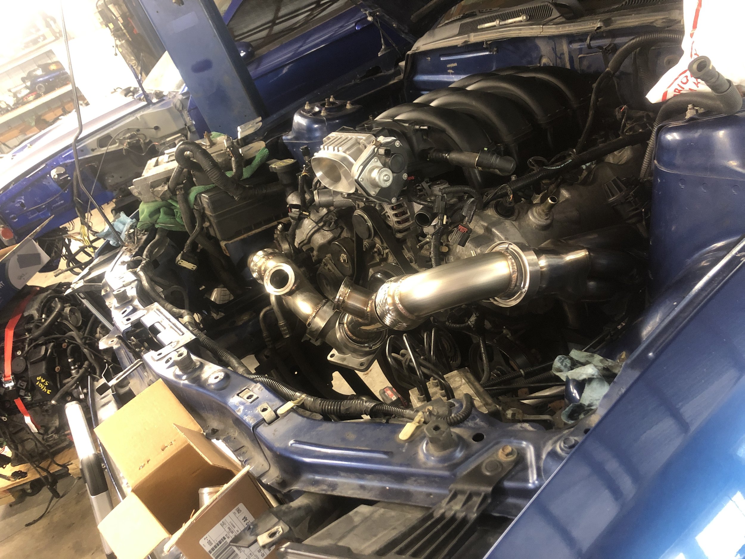 Inside the engine bay of a car, with a focus on a large, shiny metal turbocharger connected to the intake manifold by a polished metal pipe. The car's body is blue, and the engine is partially disassembled, with various hoses, wires, and components v