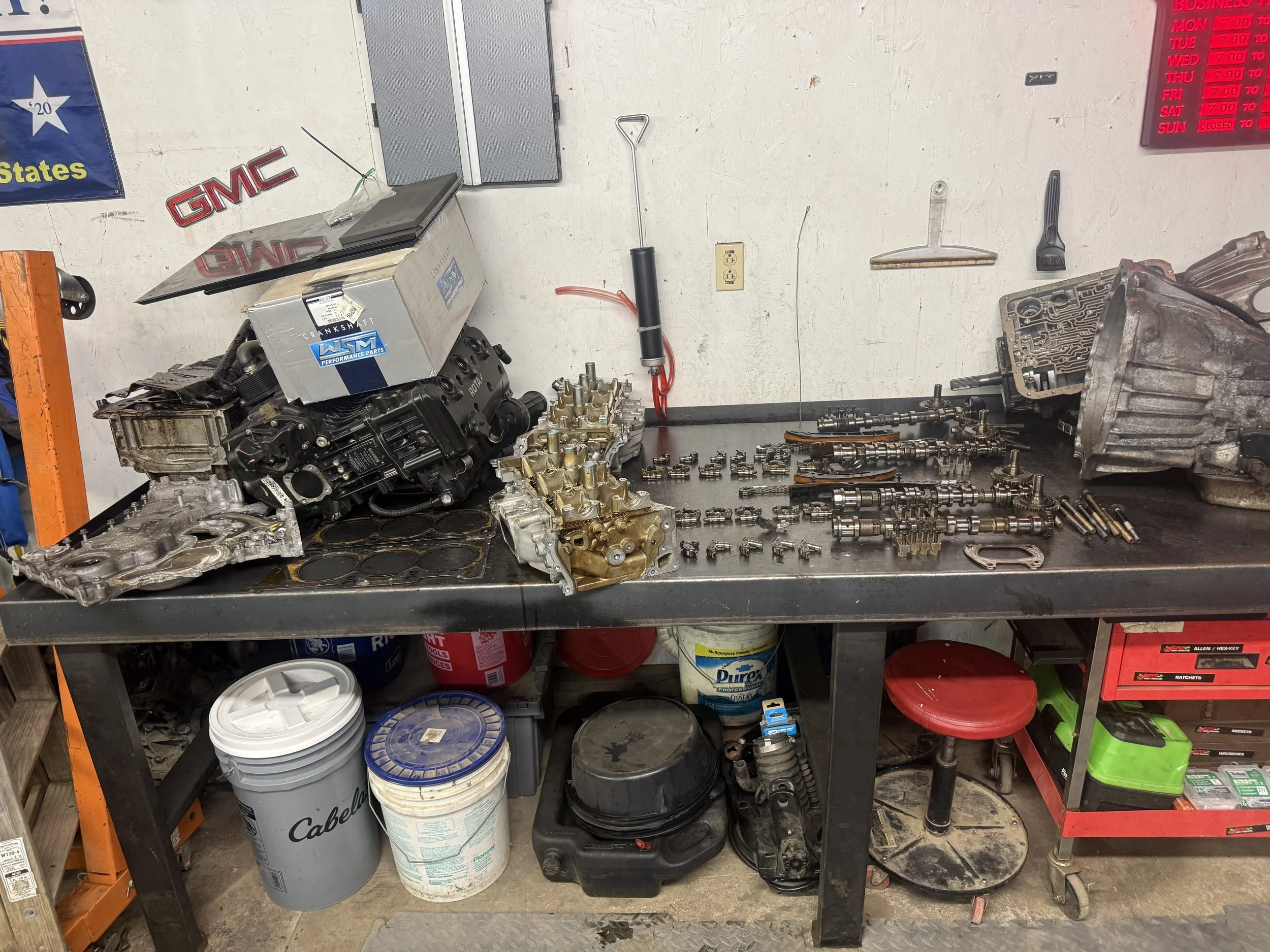Workbenches with engine parts, engine block, valves, camshafts, and related components in a garage or workshop setting.