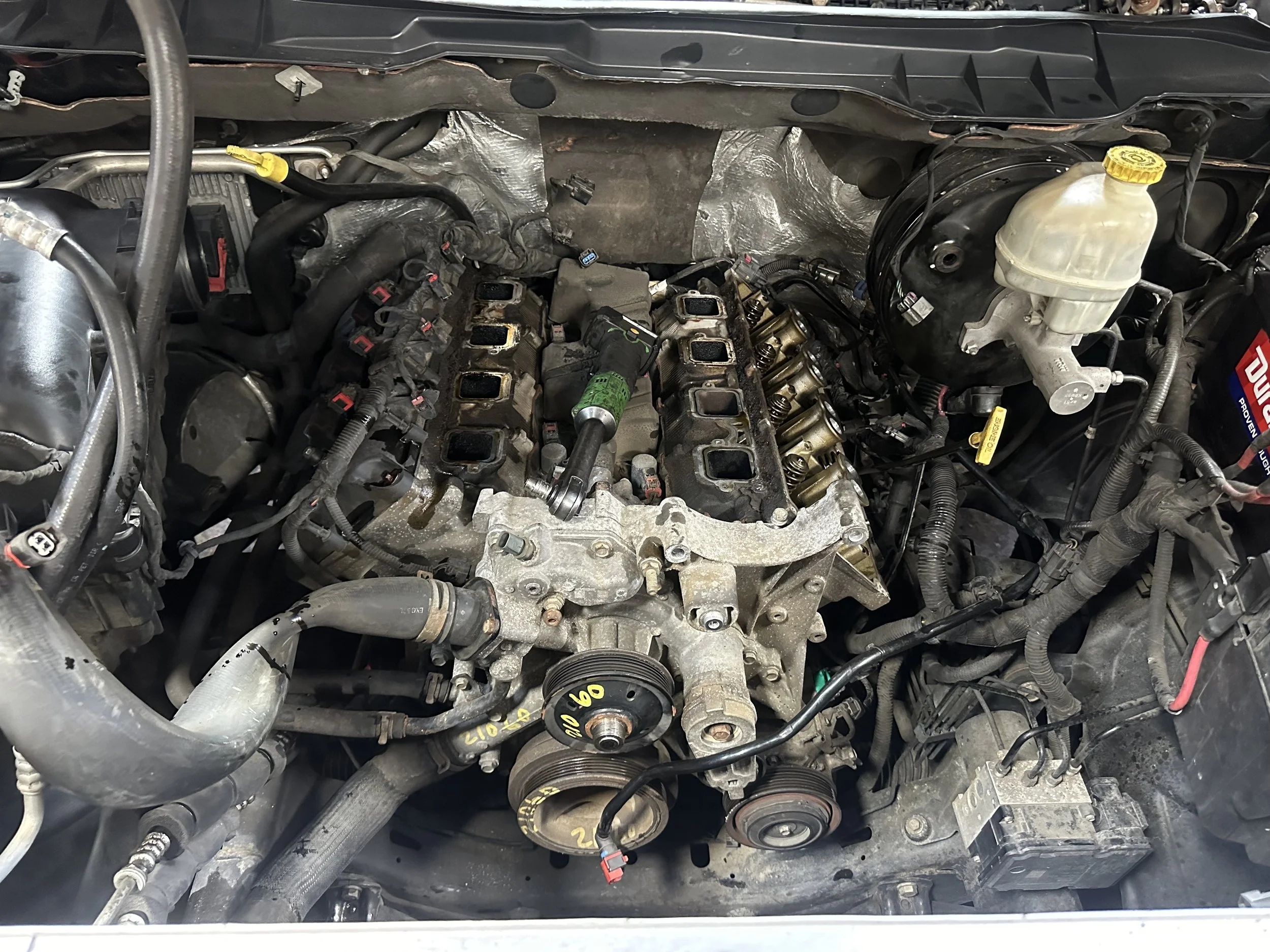 Open car hood showing engine with valve cover removed, exposing the cylinder head and valvetrain components, with various hoses and belts.