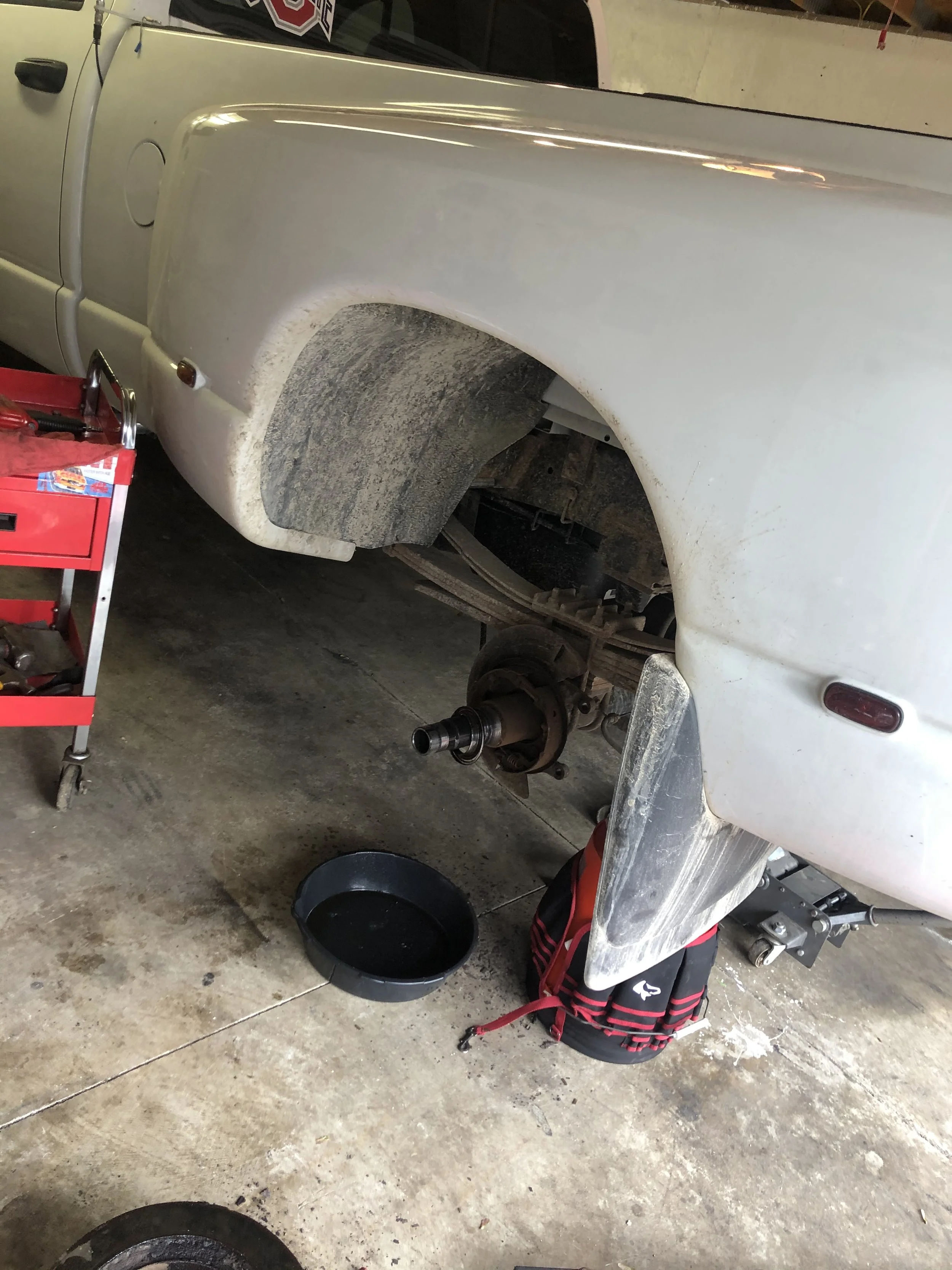 A white vehicle raised on a lift with the front wheel removed, exposing the hub and brake assembly. A red tool cart is nearby, and a black oil pan is on the floor beneath the vehicle.