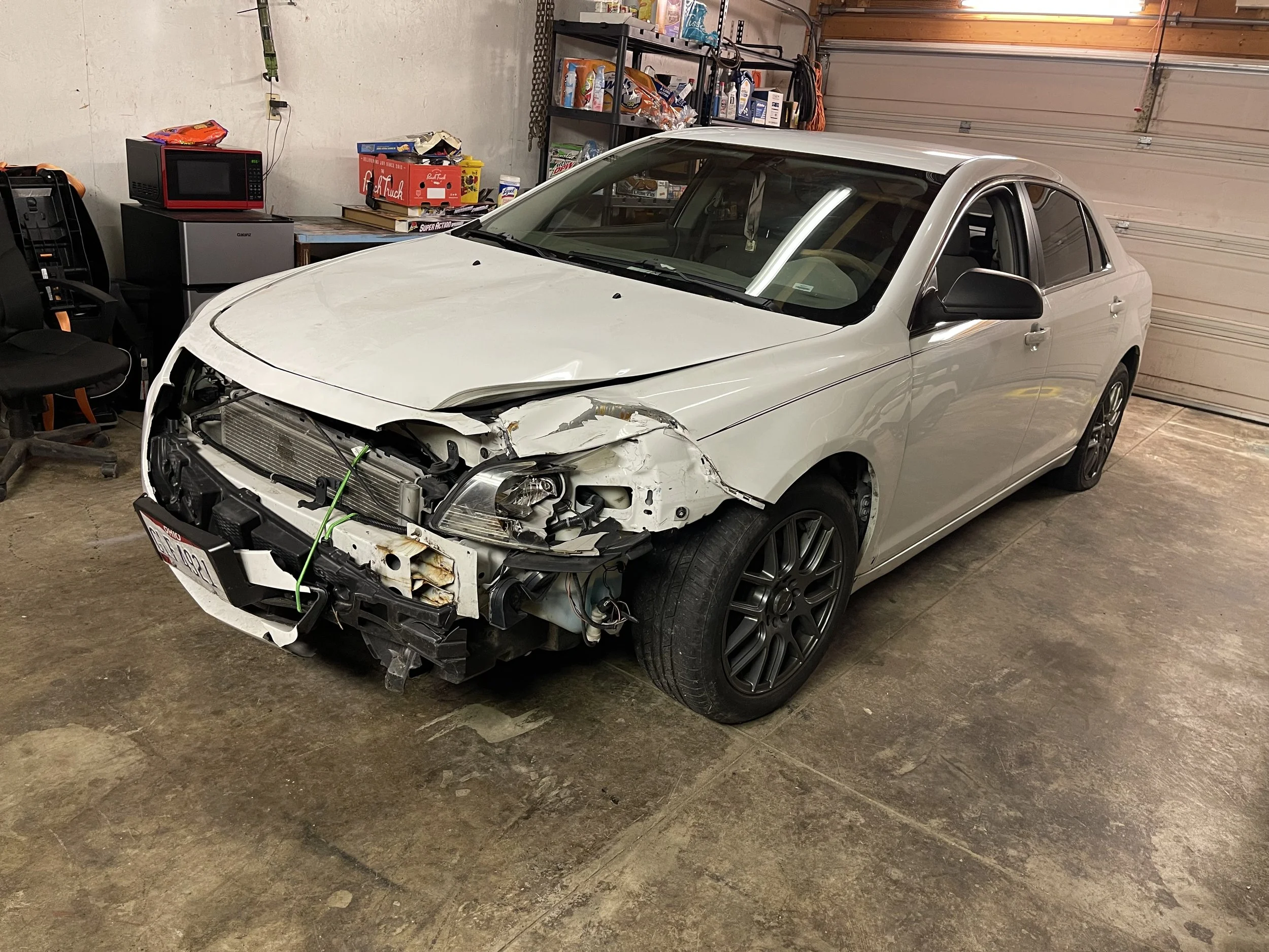 A silver sedan with front-end damage parked inside a garage. The front bumper, grille, and headlights are missing or damaged, exposing internal components. The garage has shelves with various items and a children's toy box.