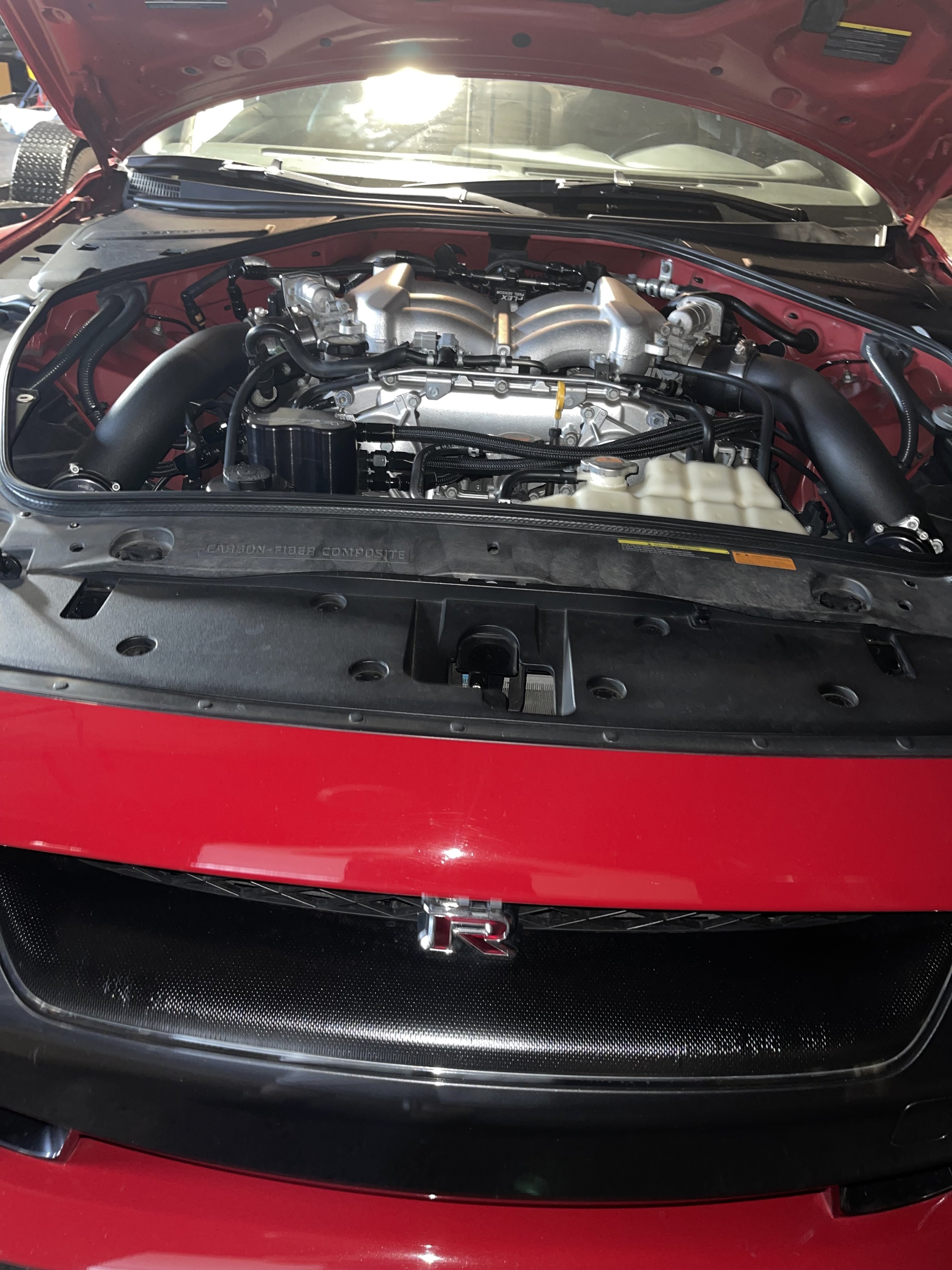 Front view of a red sports car with the hood open, revealing a detailed engine. The front grille includes a black mesh section with a chrome emblem at the center. The engine is mostly silver with black hoses and components, and the surrounding car parts are also visible.