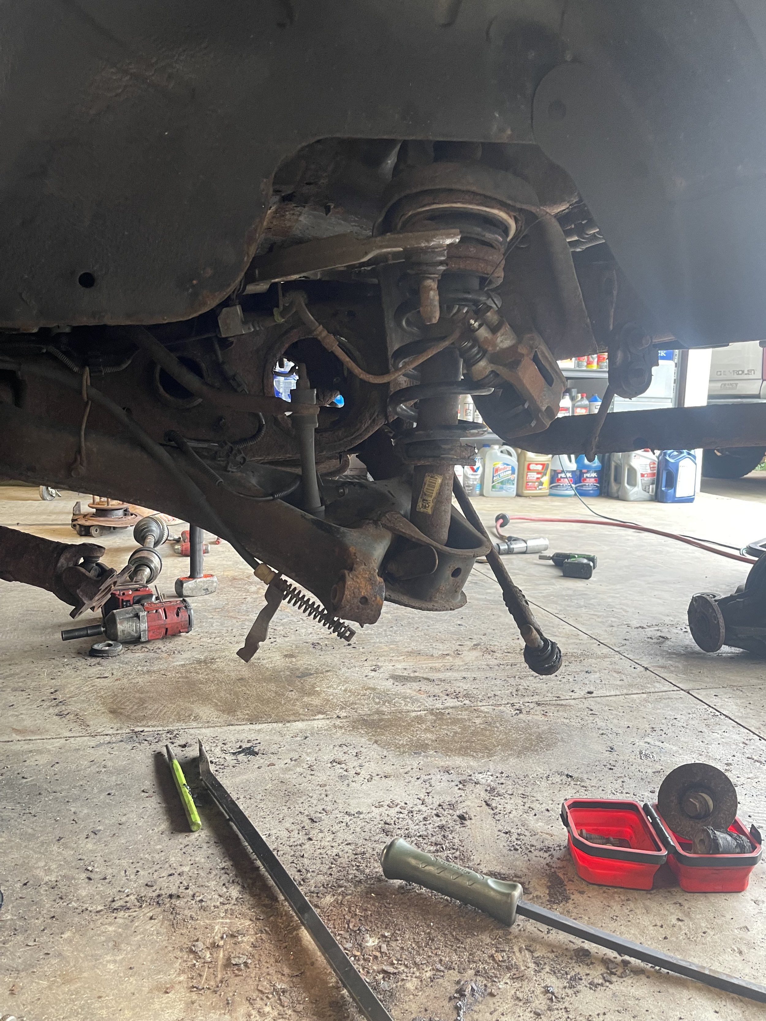 Underneath of a vehicle being worked on, with tools and parts scattered around, including a socket, a screwdriver, and a red toolbox, revealing the vehicle's suspension and undercarriage components.