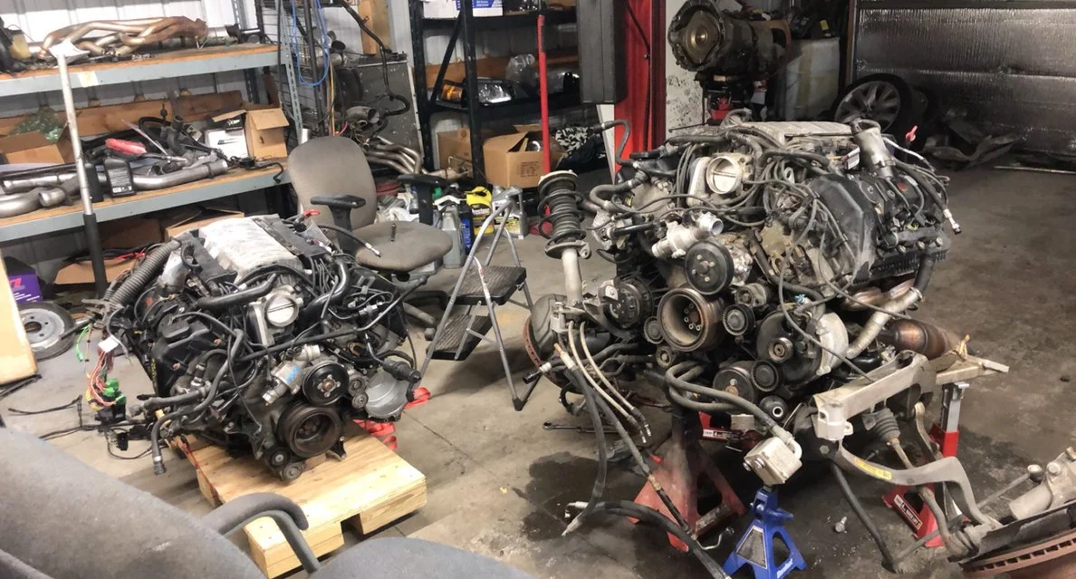 Two car engines on stands in a garage or workshop, surrounded by tools, shelves with car parts, and a chair.