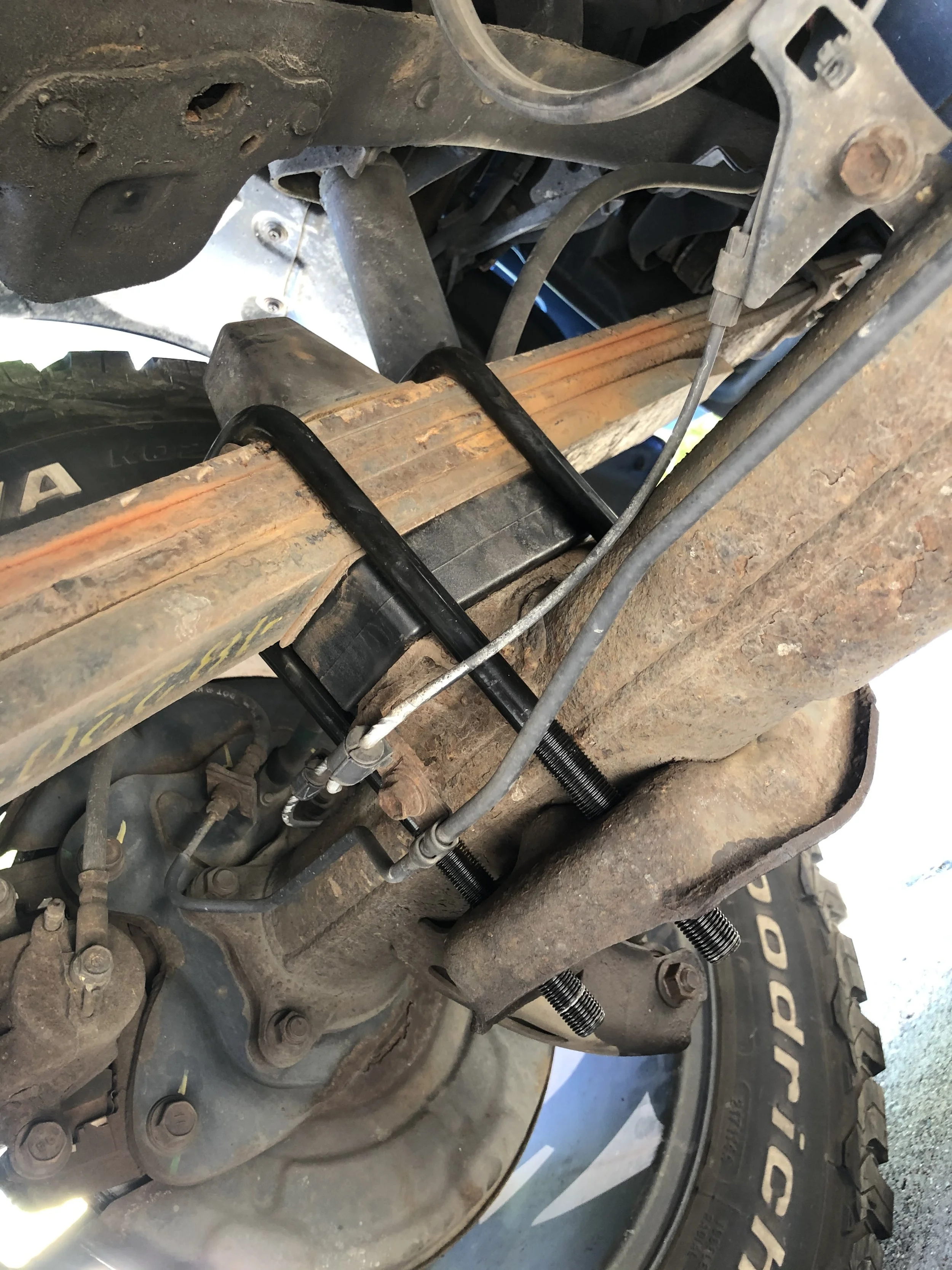 Close-up of a vehicle's steering or suspension system showing rusted metal parts, black coated control arms, and hydraulic or brake lines.
