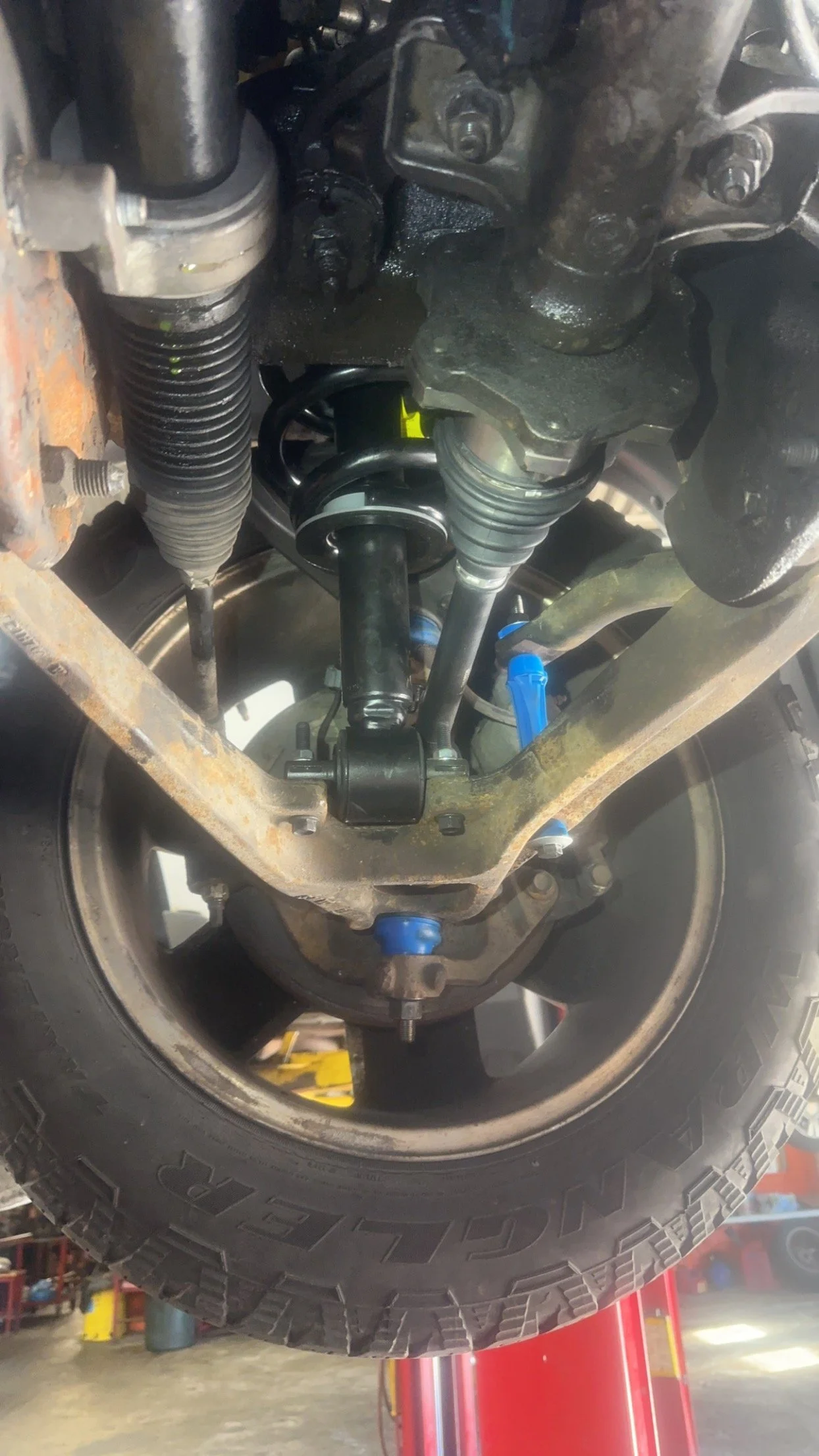View of a vehicle's front suspension, including coil spring, shock absorber, steering knuckle, and tire, with some rust on the metal parts.