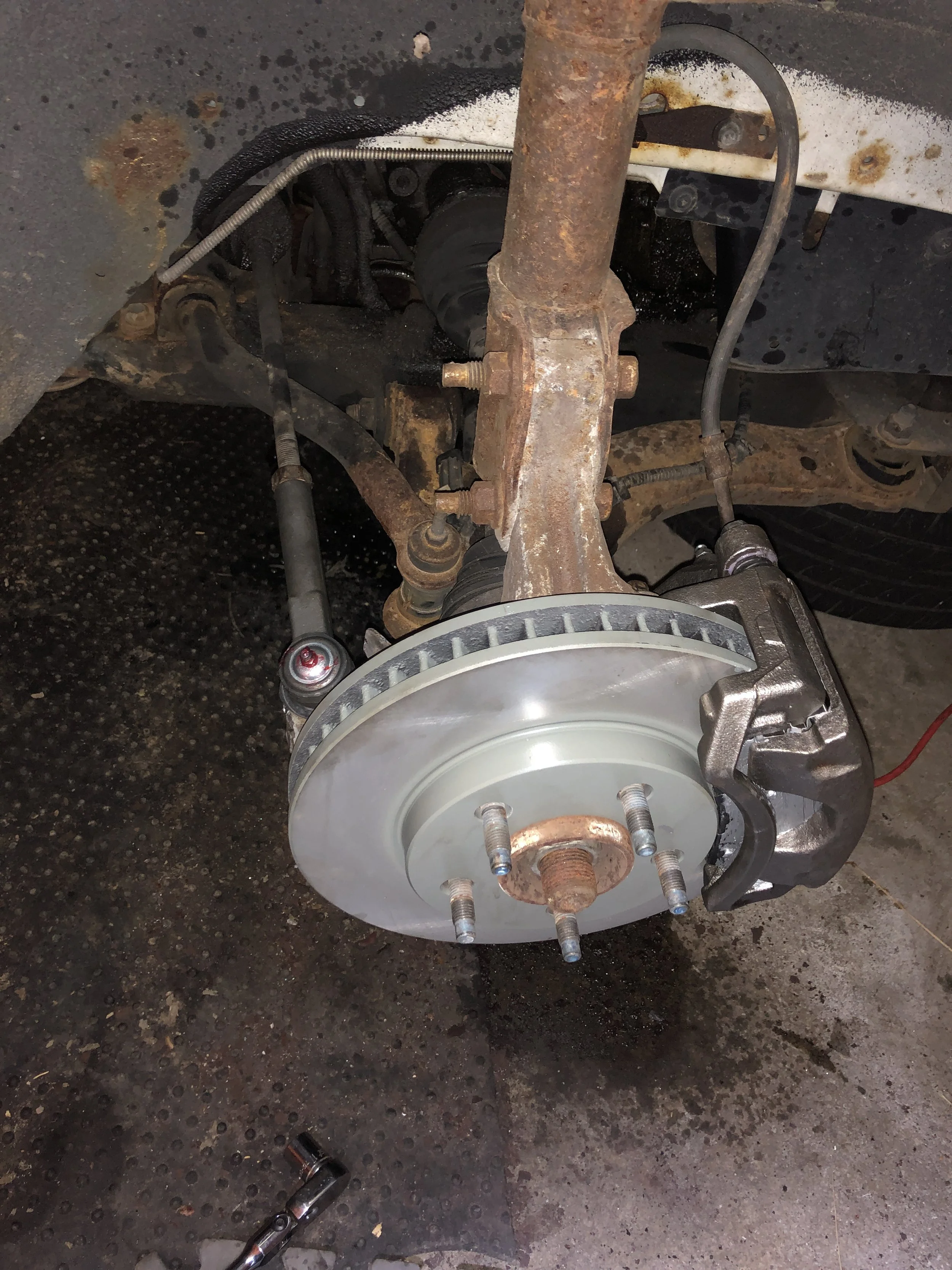 The image shows a car's front suspension and brake assembly with the wheel removed, revealing rusted and clean metal components, including the brake rotor, caliper, and suspension parts.